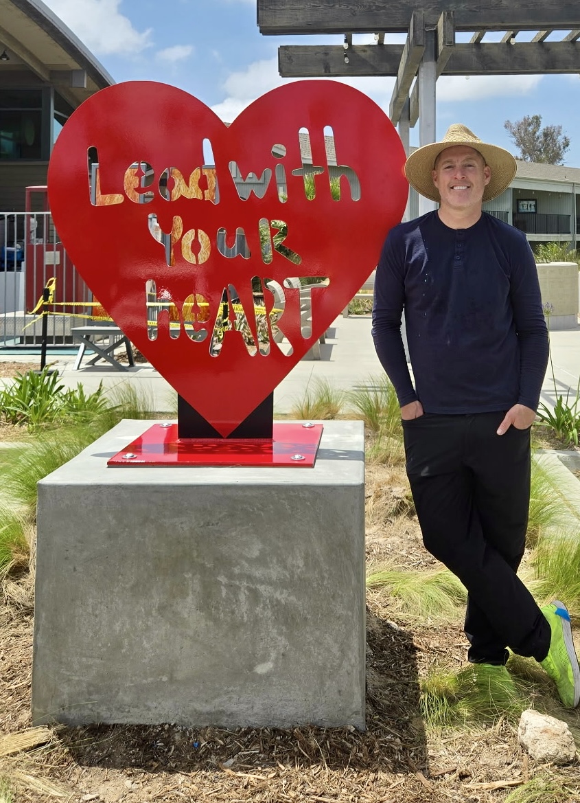 Person standing next to a large red heart sculpture with the words 'Lead with your HEART' in colorful letters, outdoors.