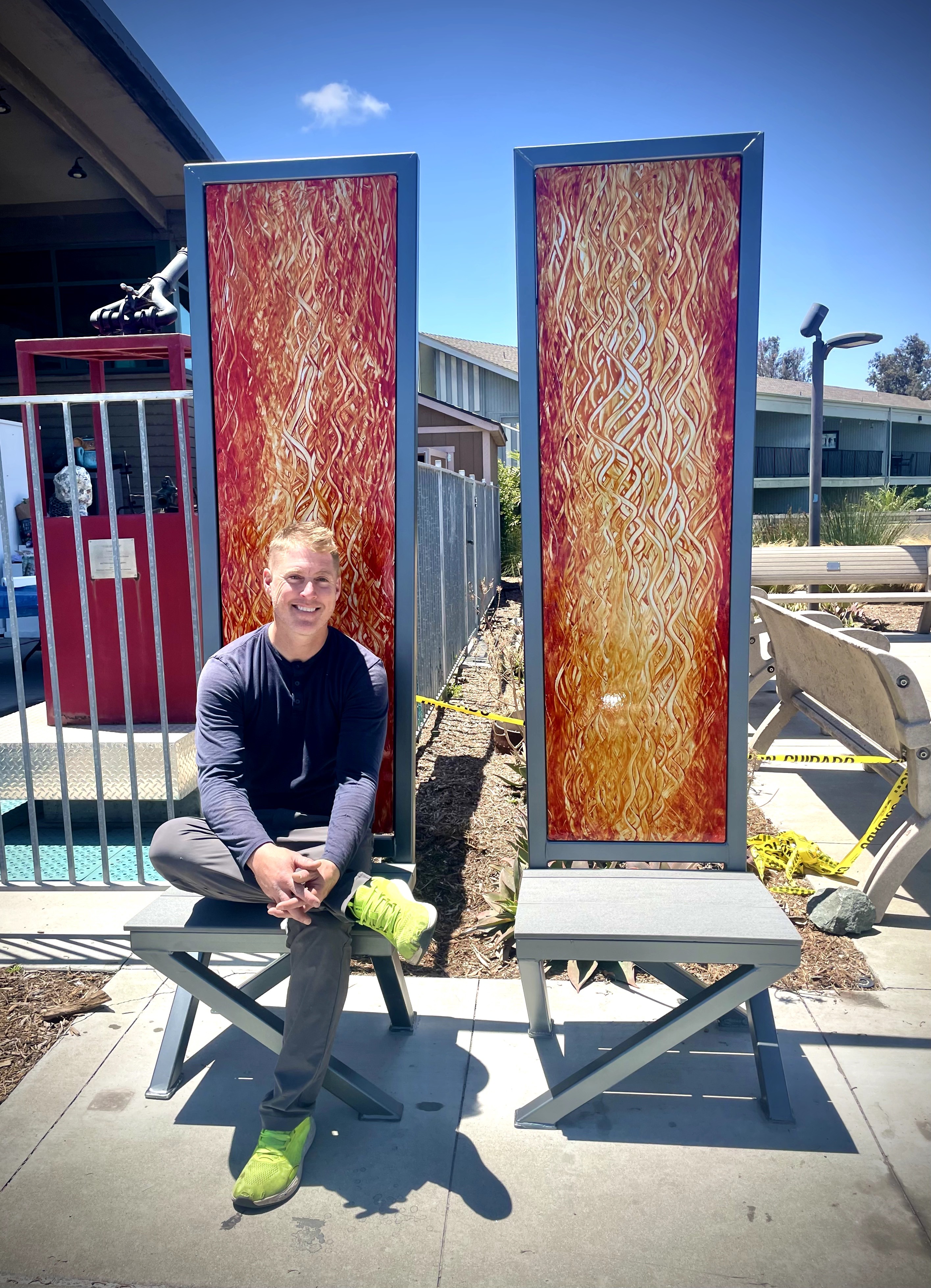 Person sitting on a bench outdoors, with two tall, narrow, fiery-colored panels behind them, under a blue sky.