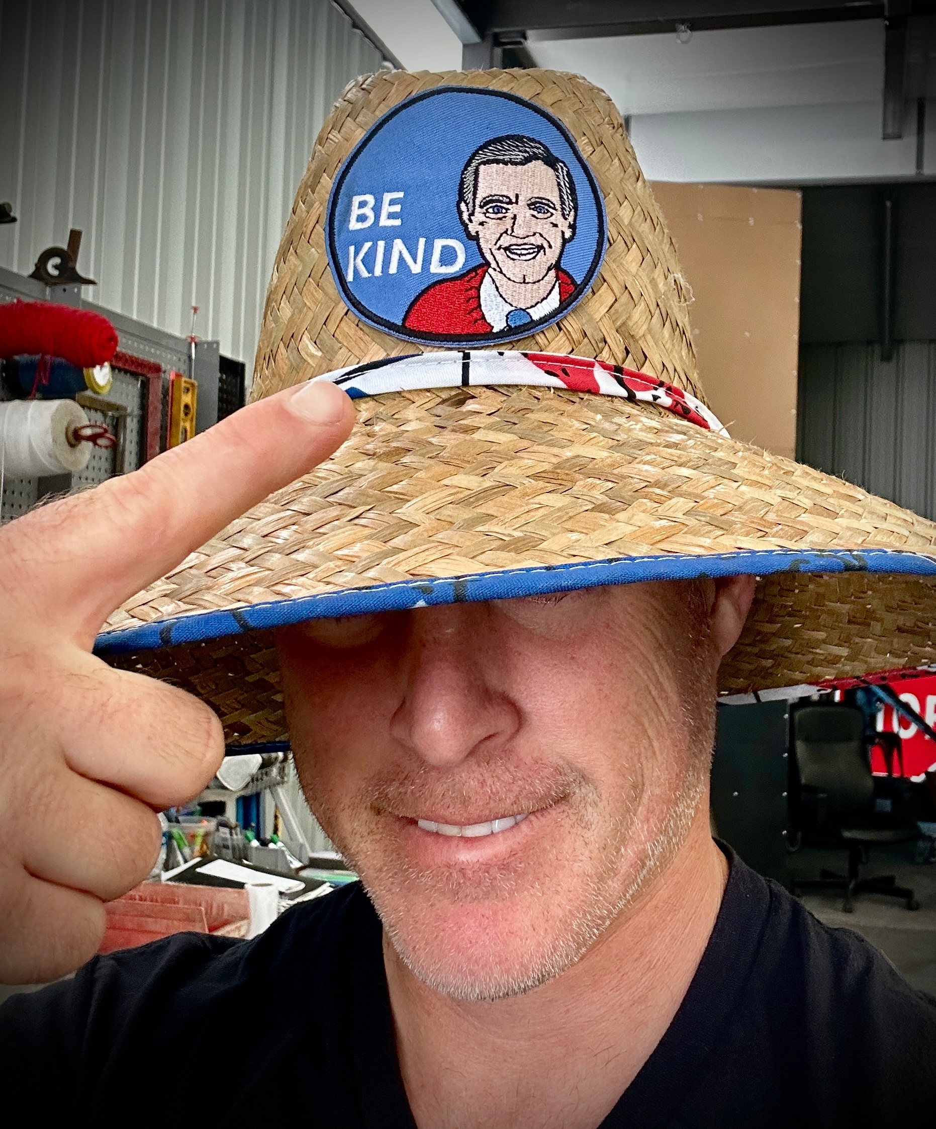 Person wearing a straw hat with a cartoon face and 'BE KIND' message, pointing to the hat's brim, smiling.