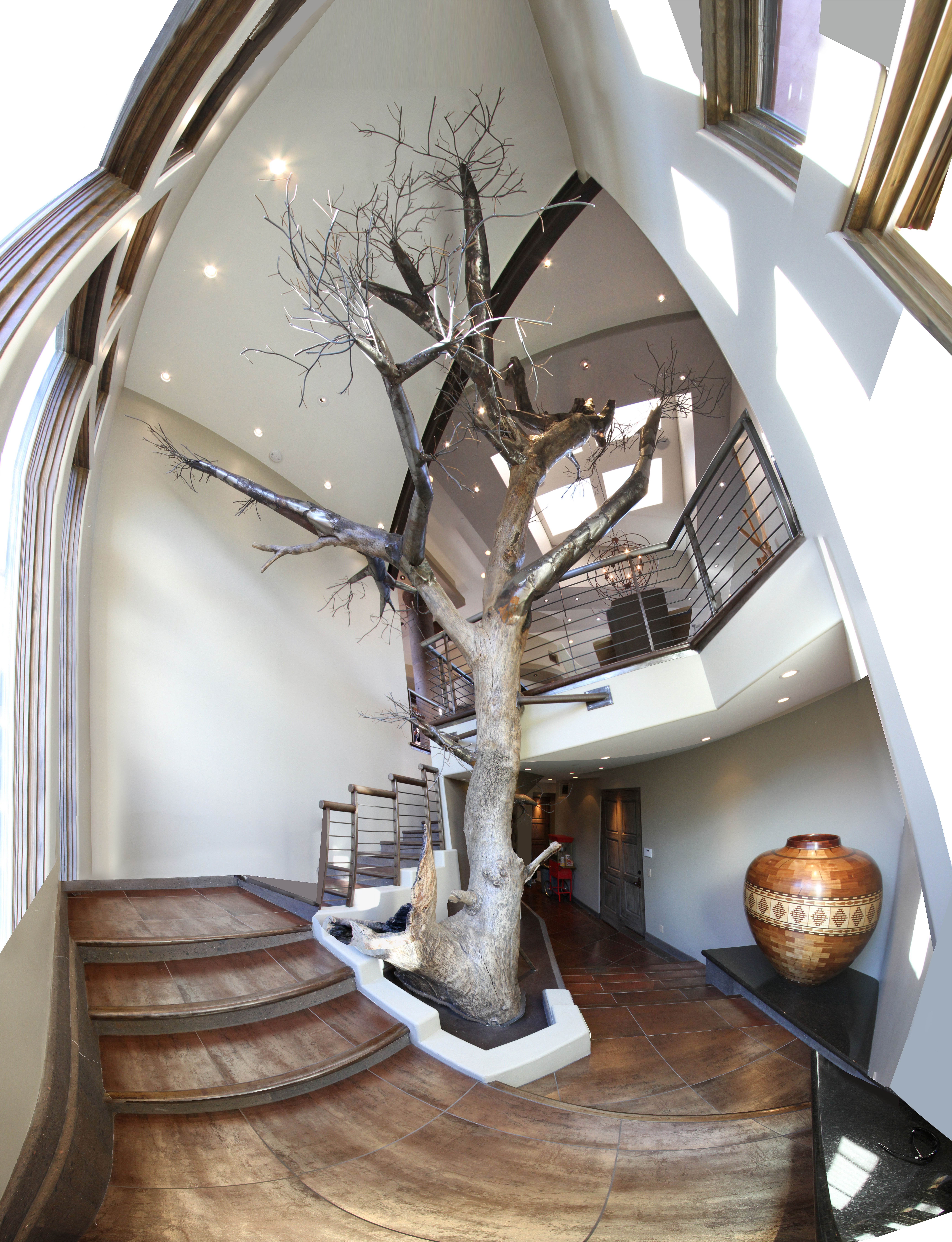 Interior view with a large leafless tree inside a building, staircase, and decorative vase, seen from below with curved ceiling.