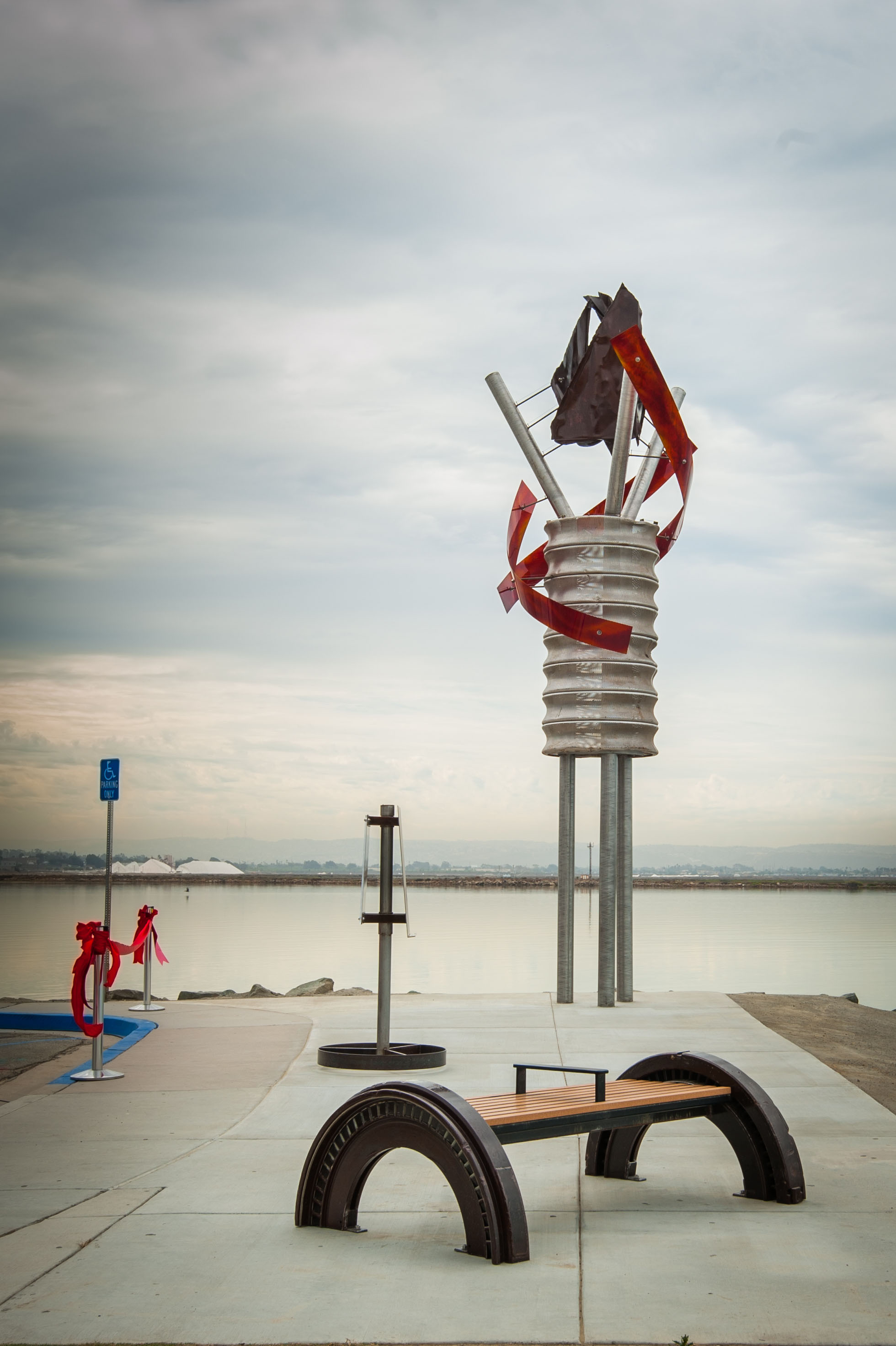 Sculpture of a horse on a coiled metal structure near water, with a bench in foreground and cloudy sky.