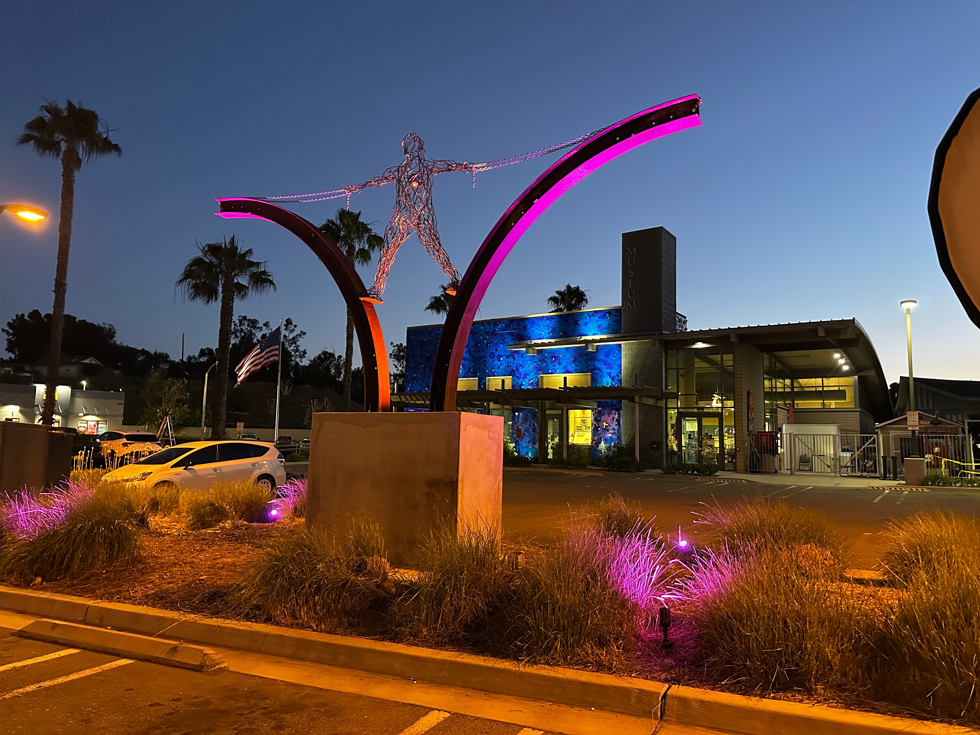 Nighttime scene with illuminated art installation, palm trees, and a building with colorful lights, pink and purple lighting on plants.
