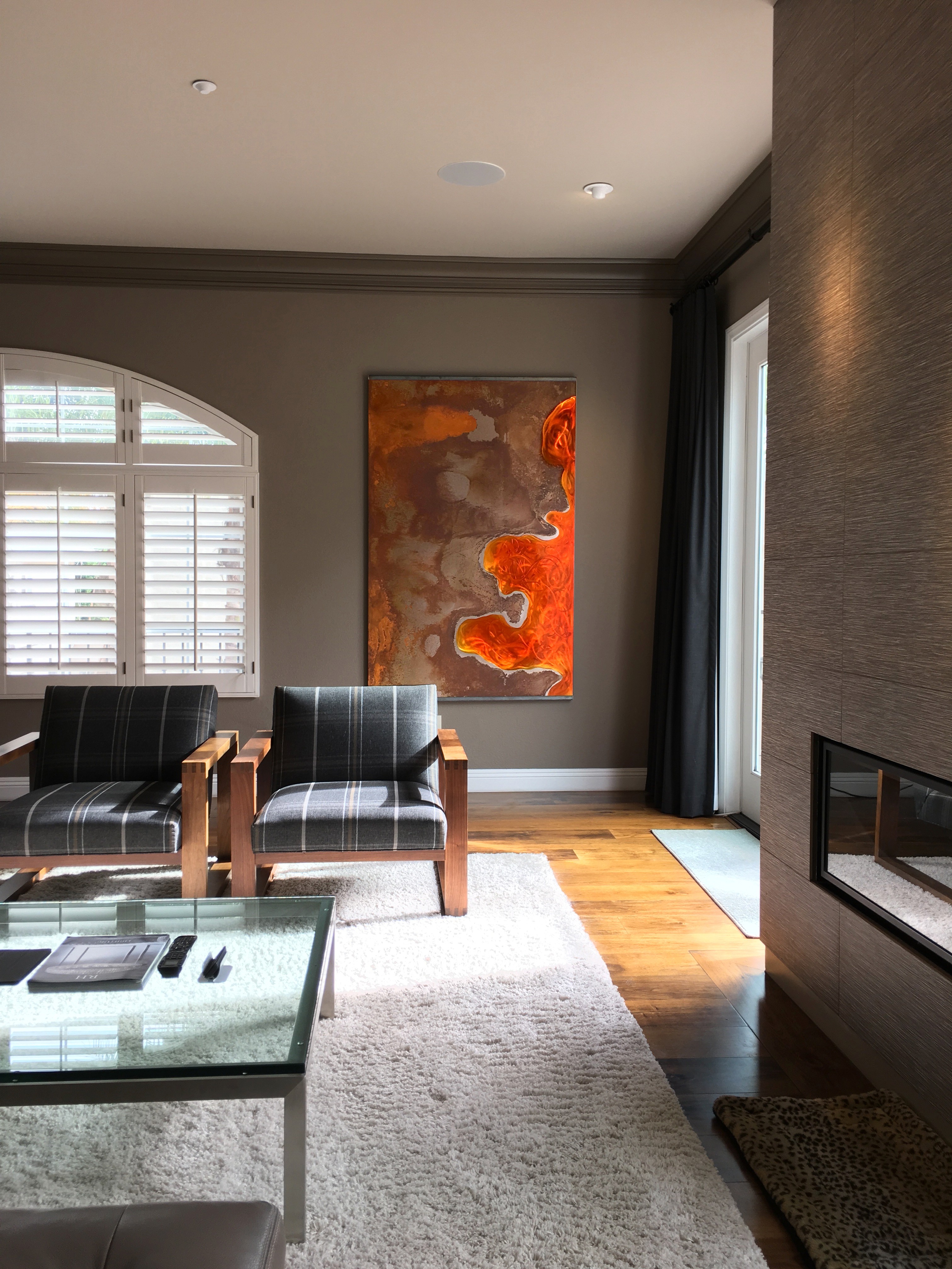 Living room with two black armchairs, a glass coffee table, a large abstract painting, and a window with blinds.