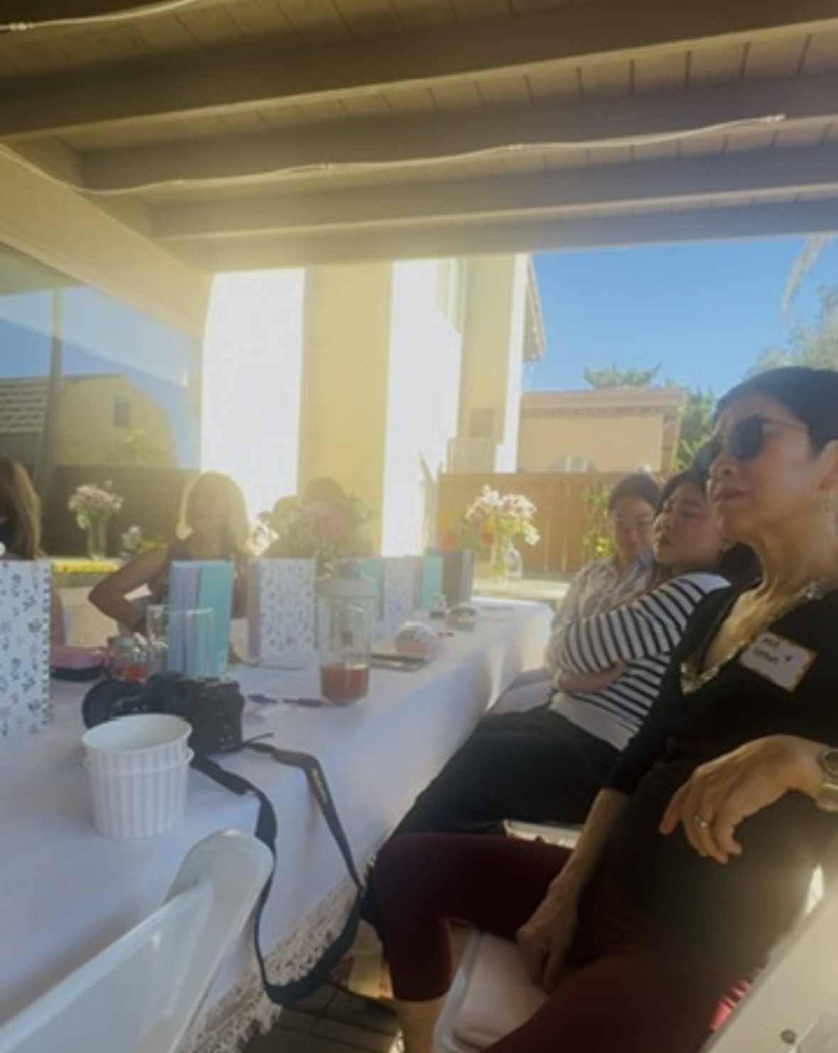 People sitting at a long table outdoors, with sunlight and a blue sky visible in the background.