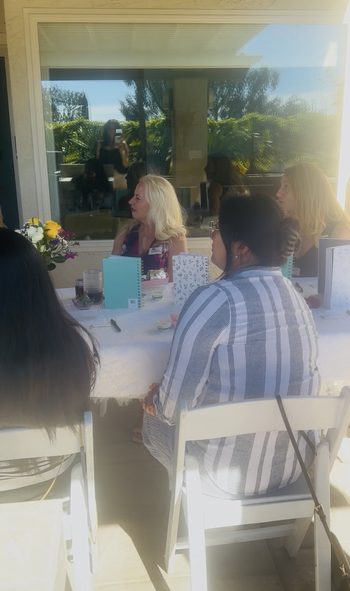 People sitting at a table outdoors, with a window reflecting trees and sky, sunlight shining, and flowers on the table.