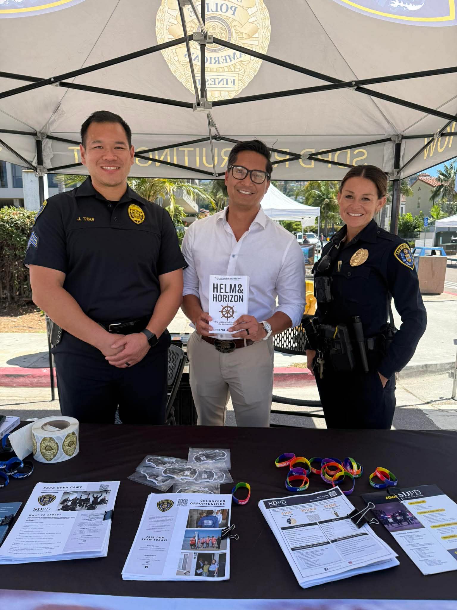 Two police officers and a man holding an award stand under a canopy, with a table of flyers and wristbands in front.