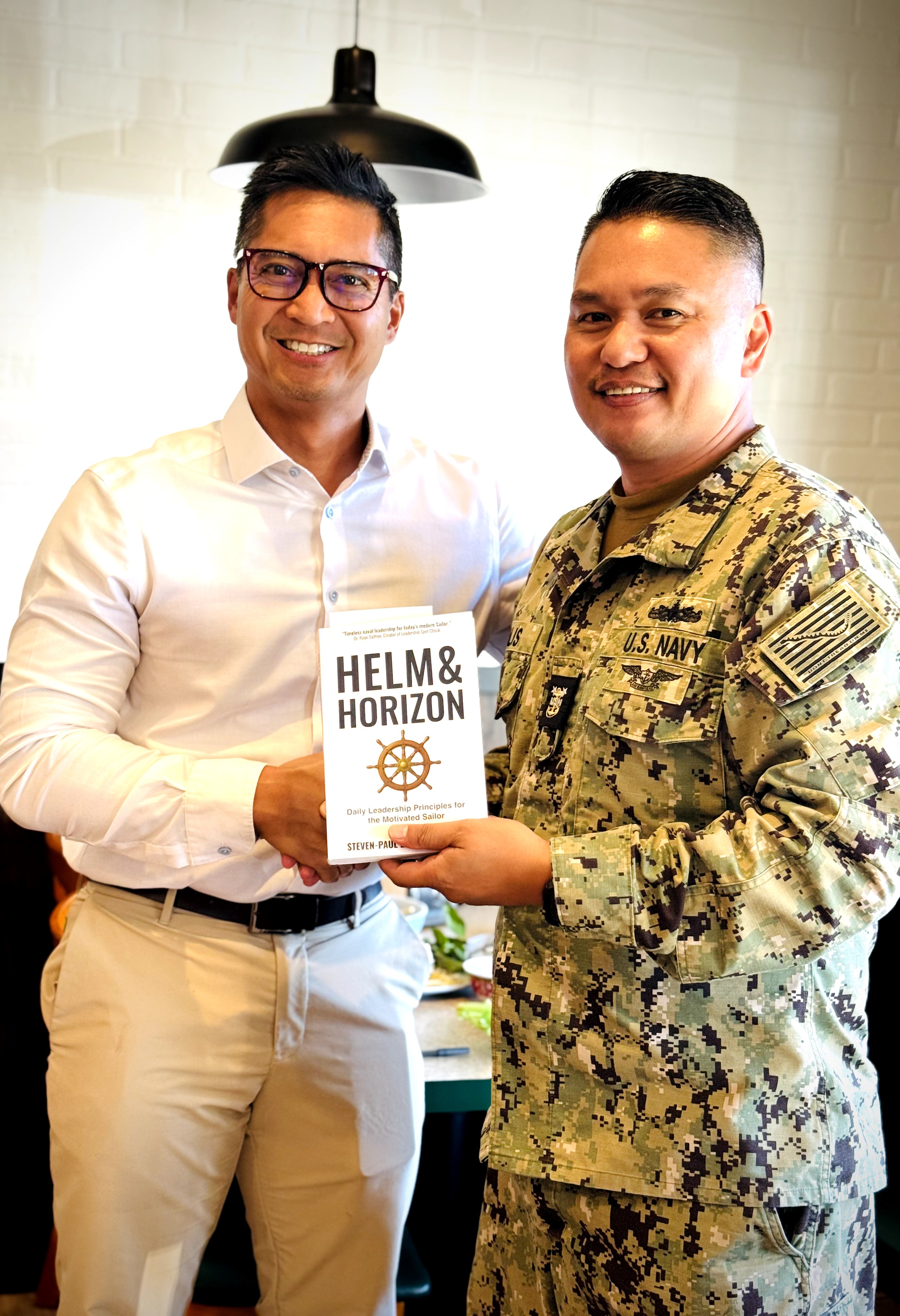 Two men, one in a white shirt and the other in military uniform, hold a book titled 'Helm & Horizon' together.