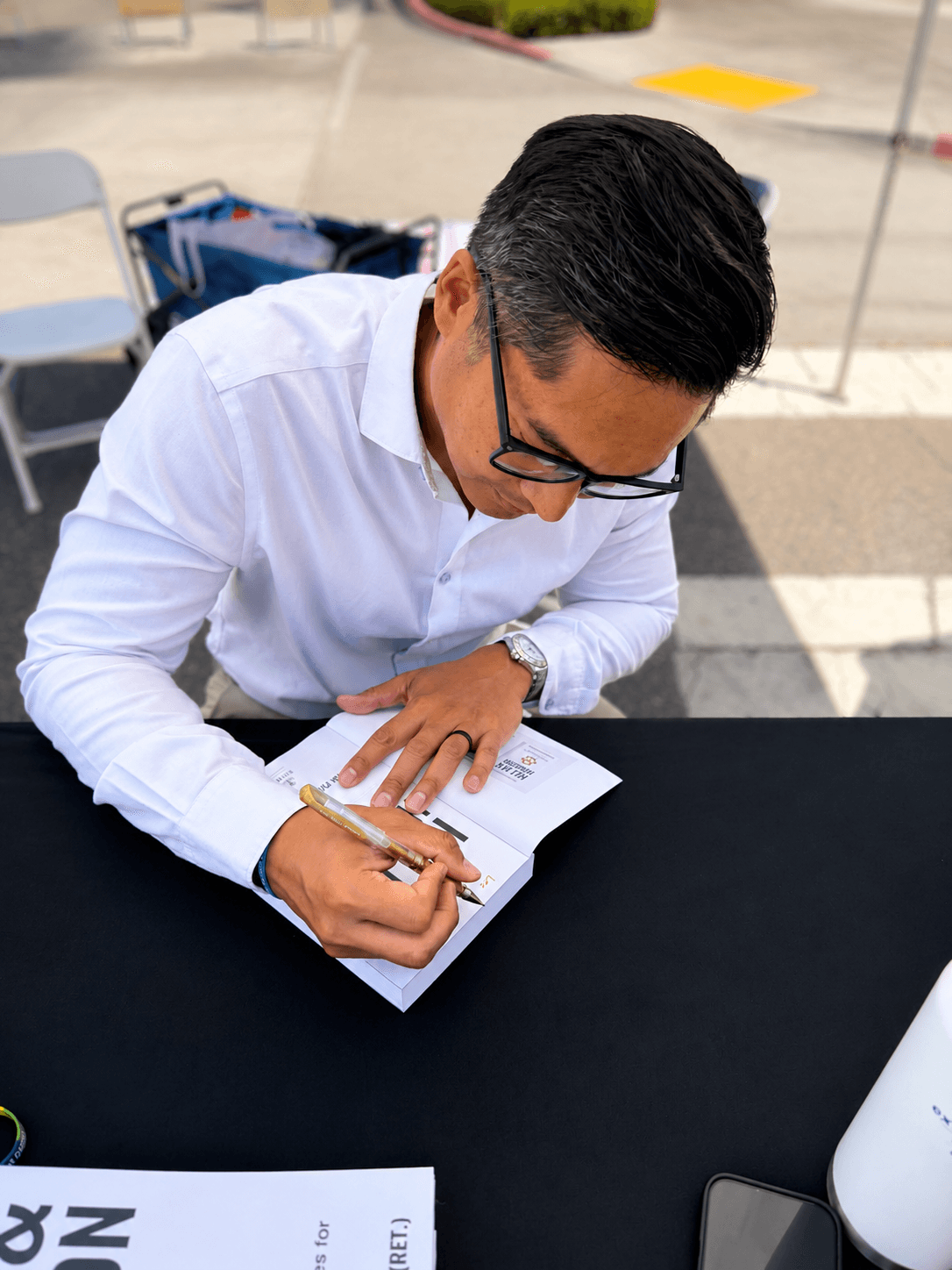 Person with black hair and glasses writing in a notebook at a table outdoors.