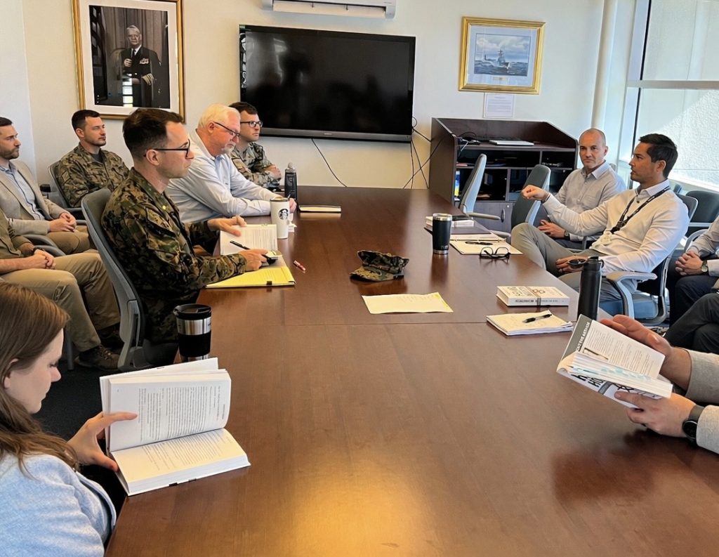 Group of people seated around a conference table in a meeting room, some reading documents, with a large screen on the wall.