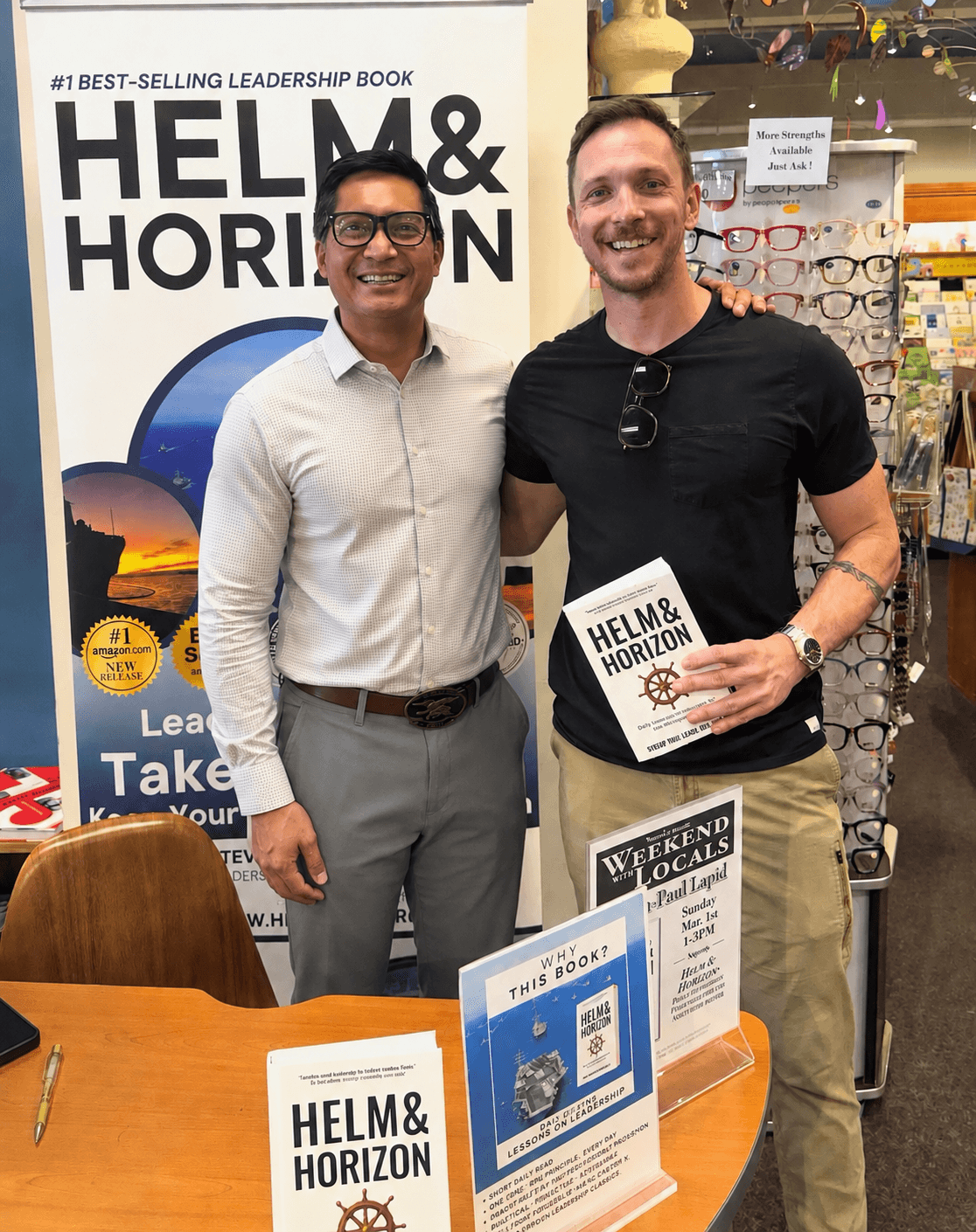 Two men standing indoors, one holding a book titled 'Helm & Horizon,' smiling, with a large book display behind them.