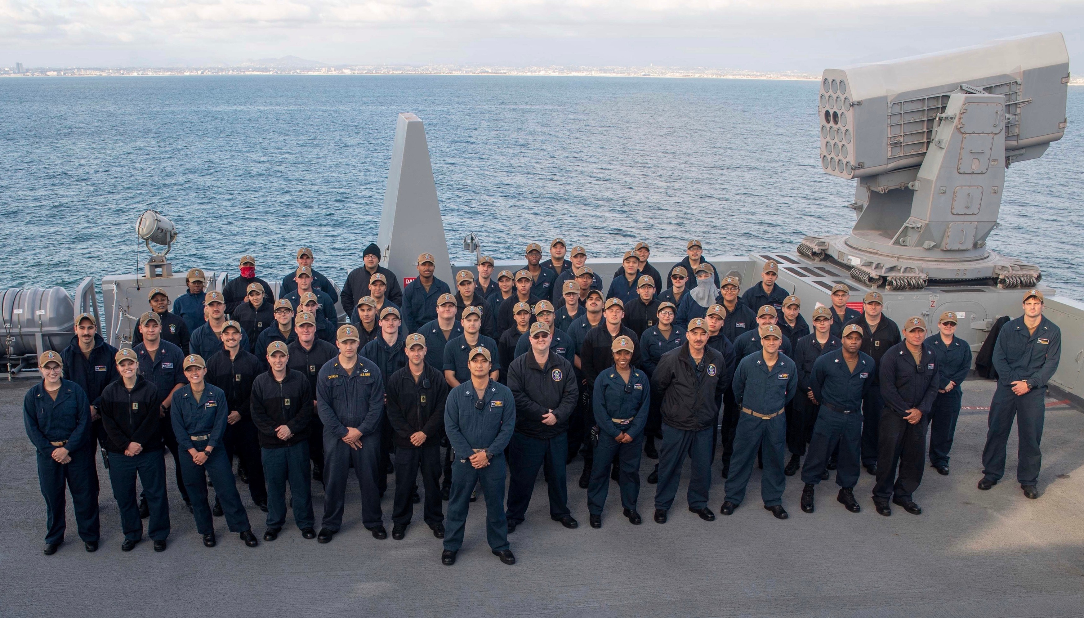 Group of people in uniform standing on a ship's deck with a large gun and ocean in background.