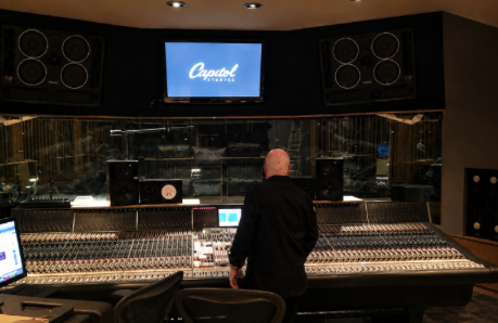 Person in a recording studio control room with mixing console and speakers, monitor screen displaying 'Capitol'.