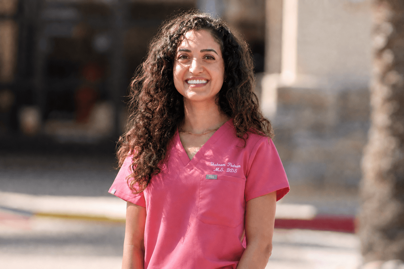 Young woman with curly hair smiling, wearing pink scrubs, standing outdoors in sunlight.