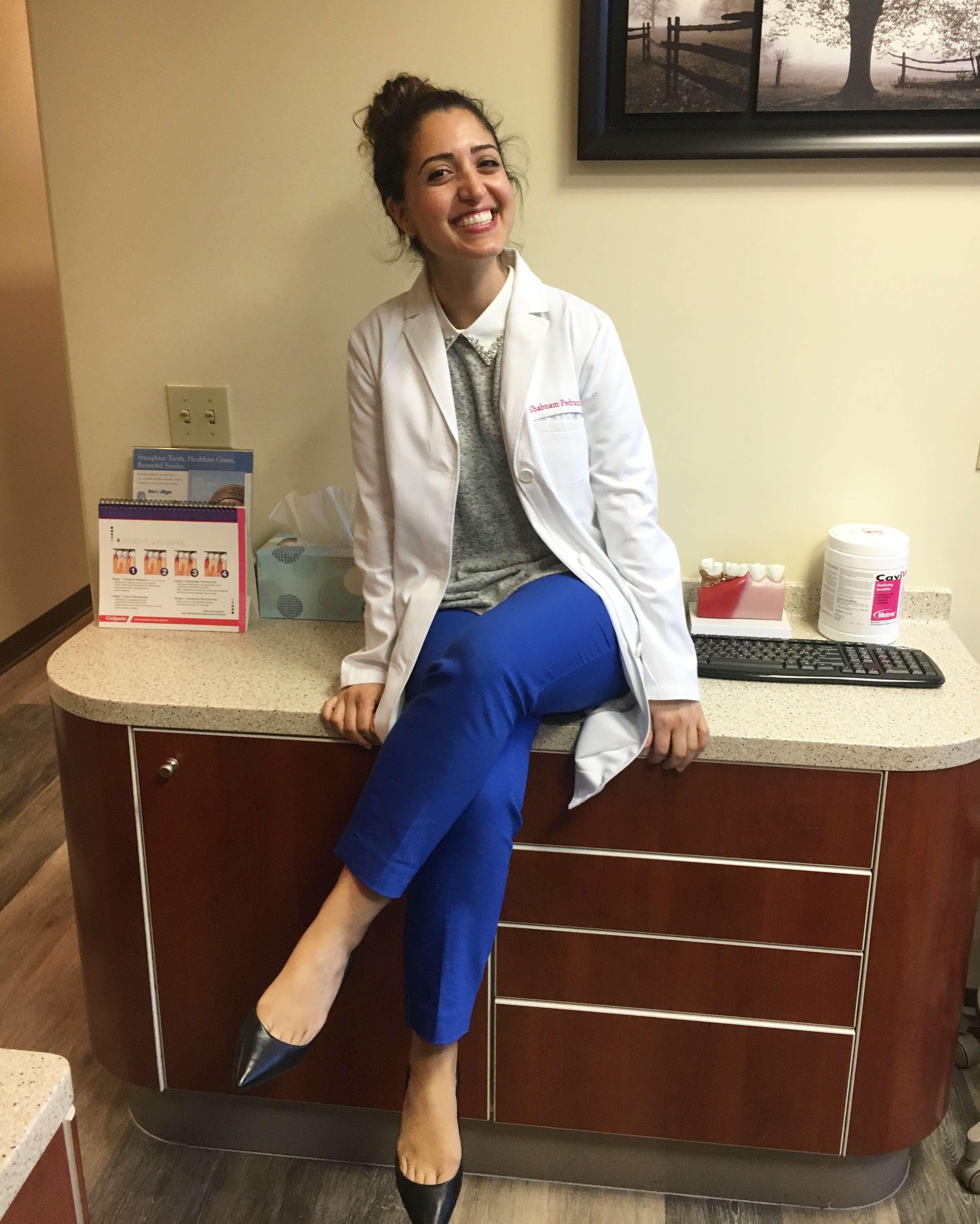 Woman in white coat sitting on a medical counter, smiling, with a computer keyboard and medical supplies nearby.