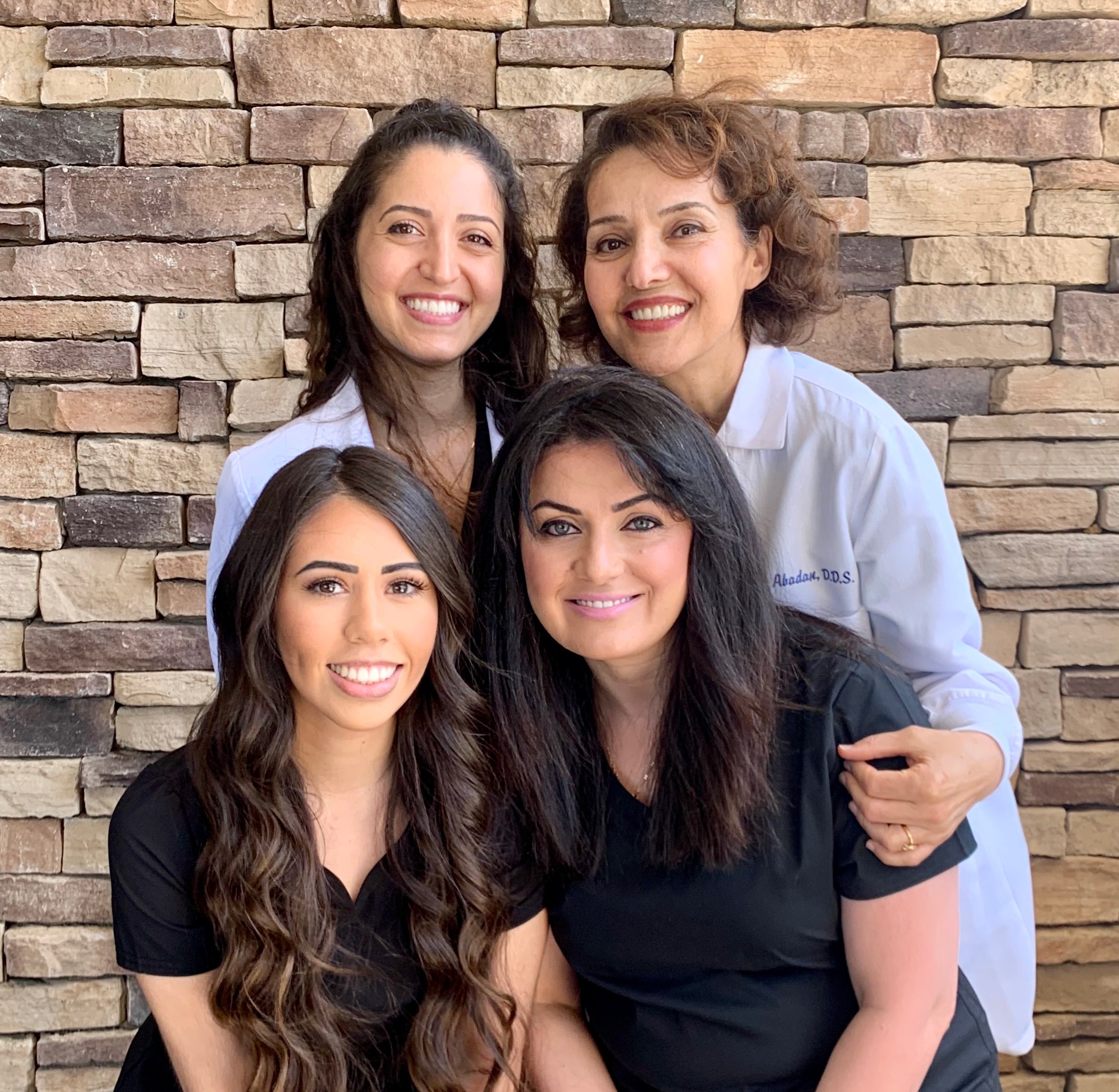 Four women smiling, standing in front of a stone wall, two in black scrubs and two in white coats.