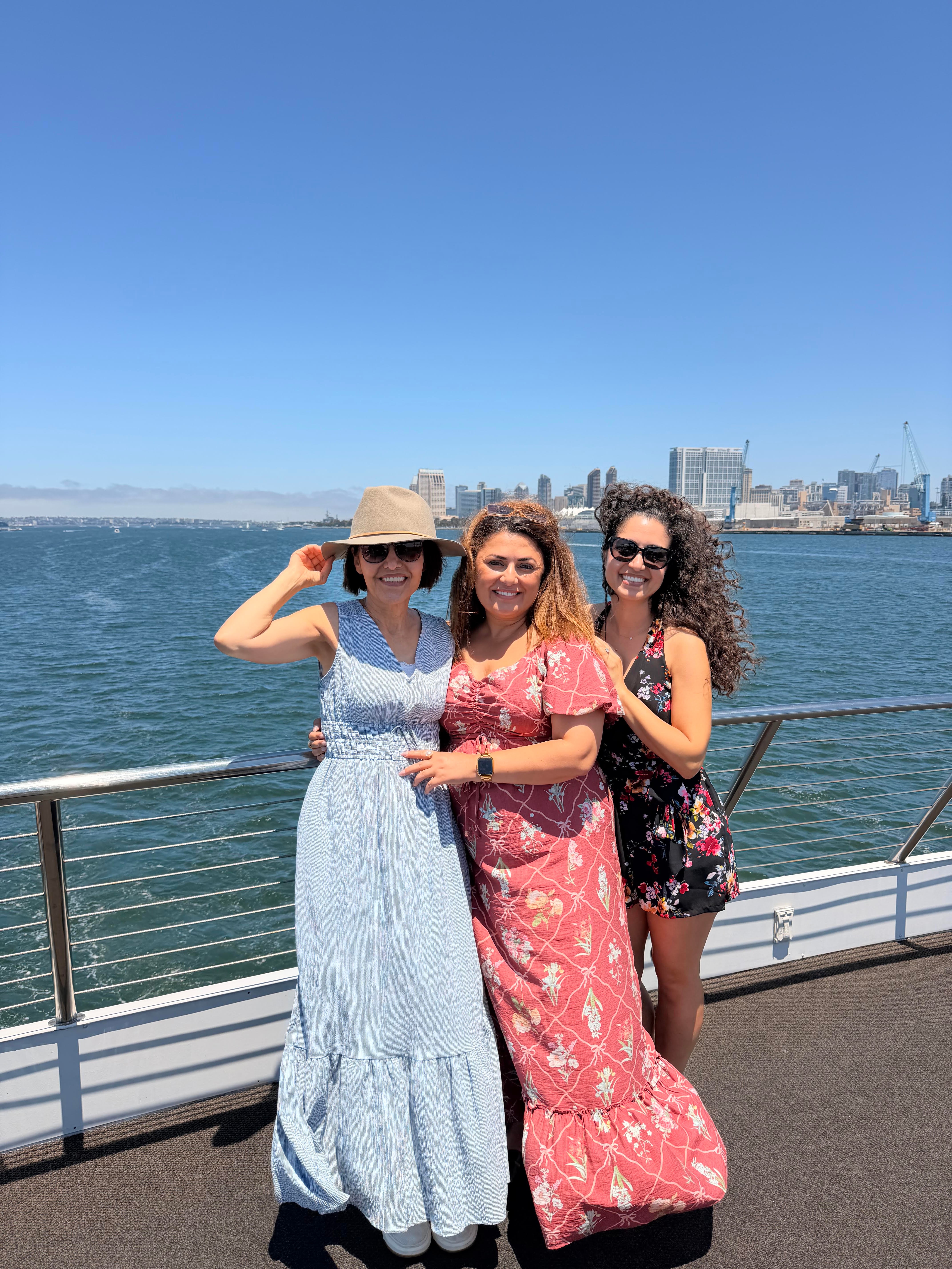 Three women standing by a railing near water with a city skyline in the background, smiling, one wearing a hat, one in a pink dress, one in a floral outfit.