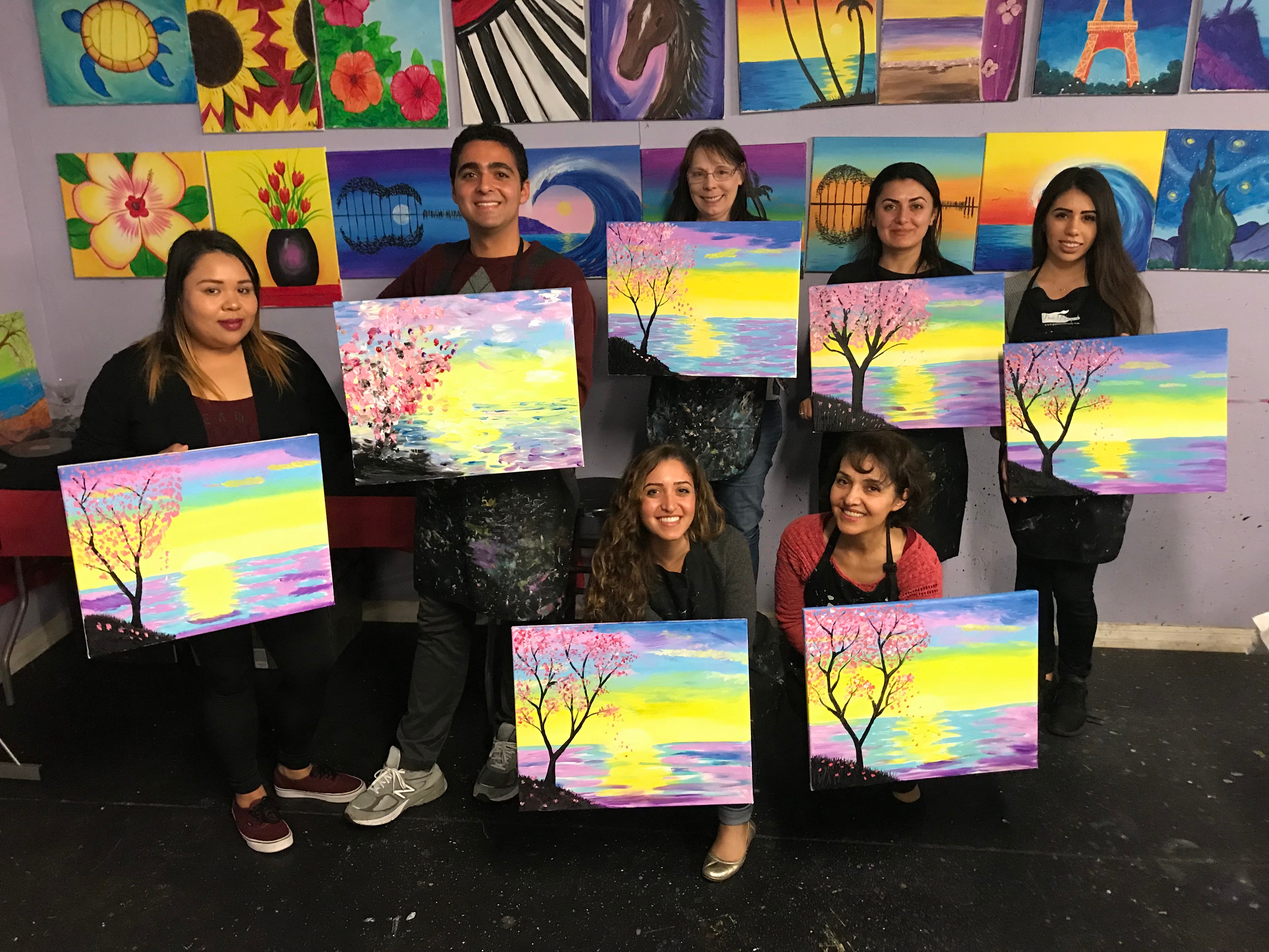 Group of nine people holding colorful landscape paintings with trees and sunsets, standing in front of a wall with various artwork.
