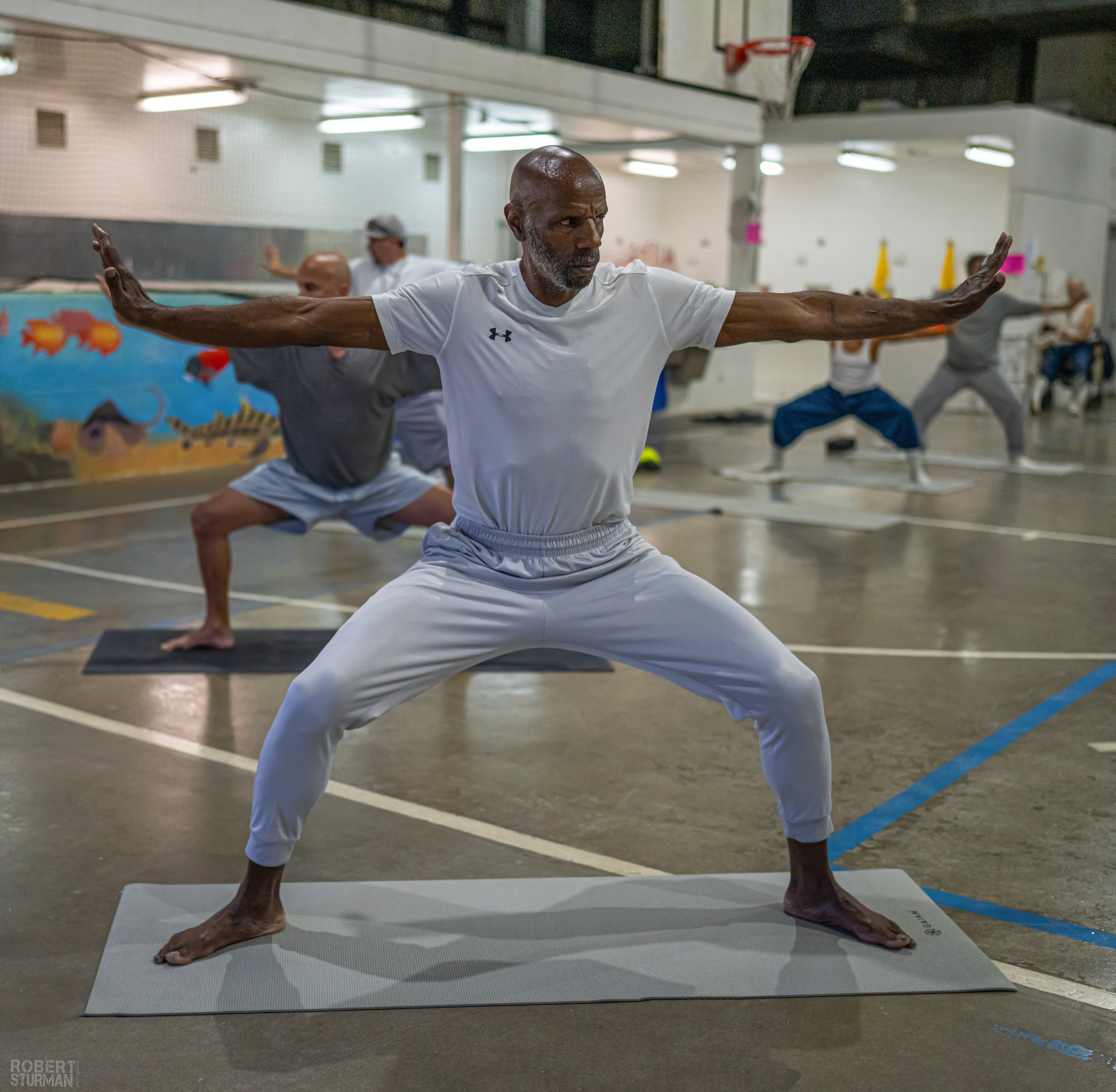 Man practicing yoga in a gymnasium with others in the background, arms extended, standing on a mat.