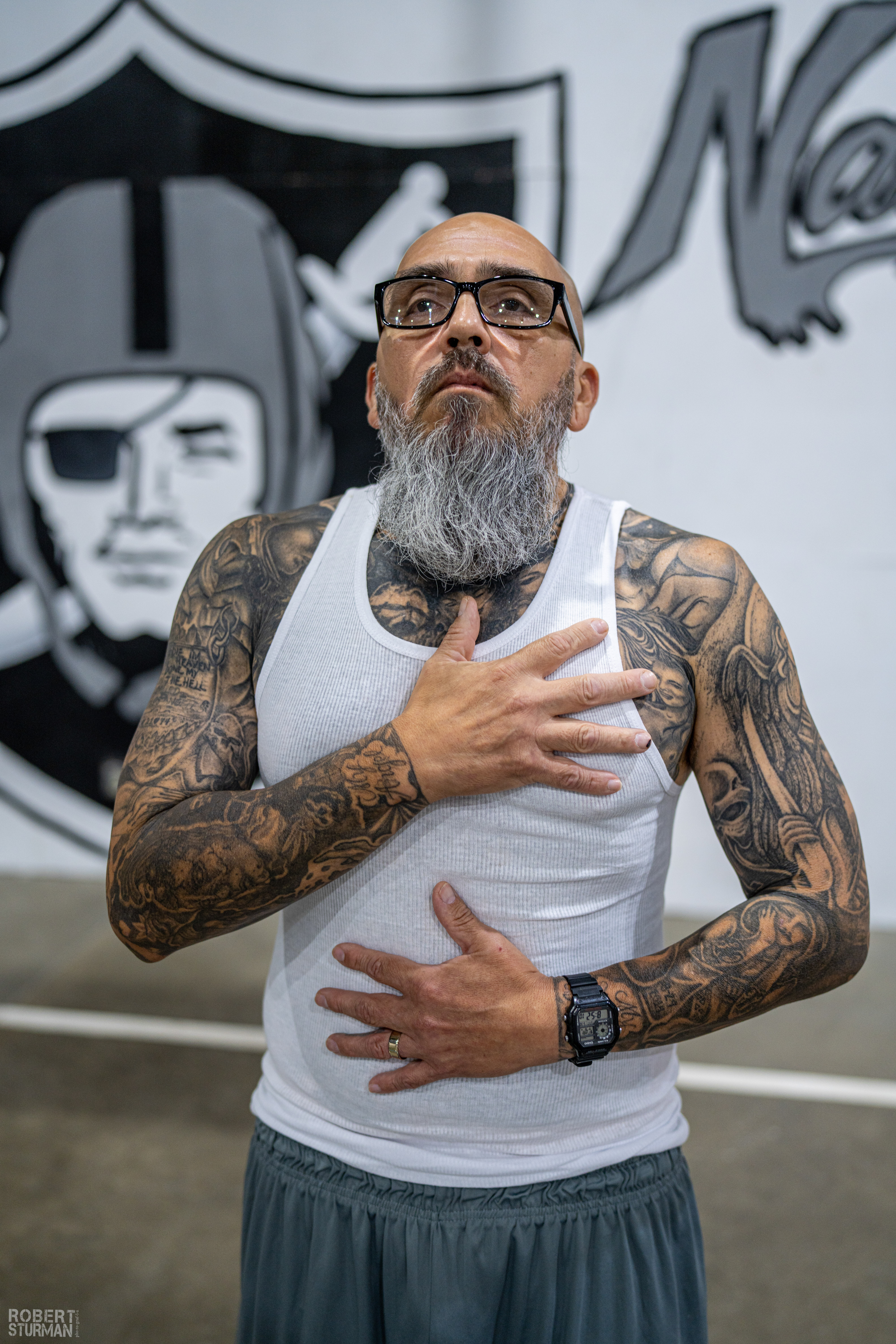 Man with glasses and tattoos on arms and chest, wearing a white tank top, standing indoors with graffiti background.