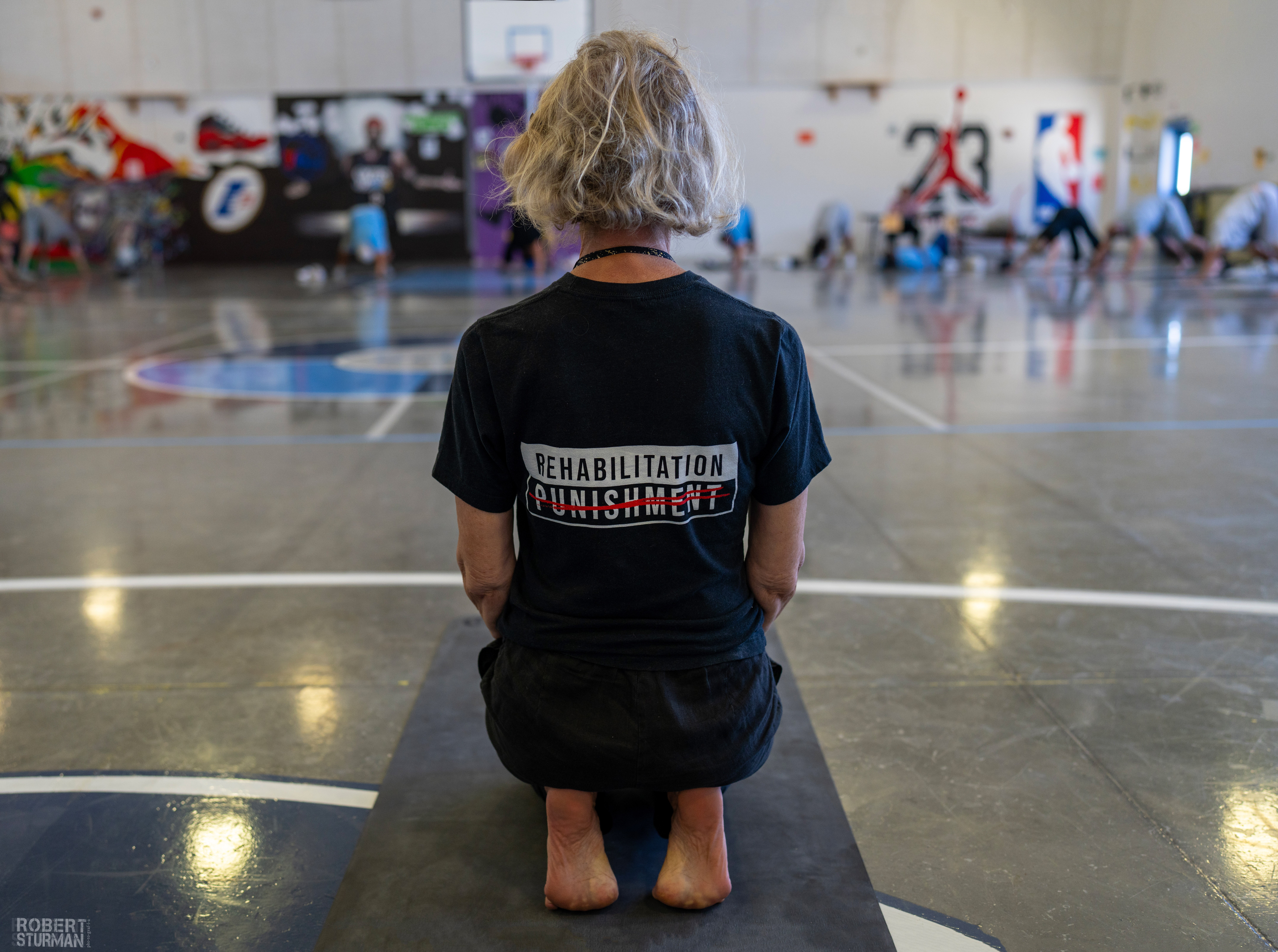 Person kneeling on a mat in a gymnasium, facing away, with a t-shirt that reads 'Rehabilitation Punishment'.