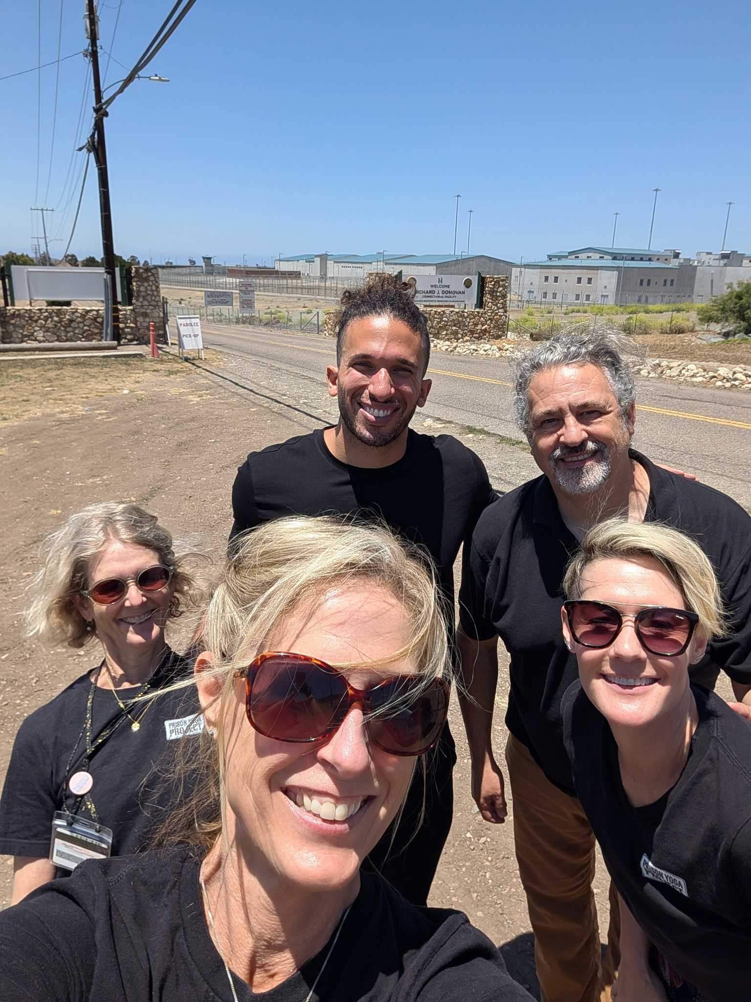 Group of five people outdoors smiling, sunny day, background with buildings and power lines.