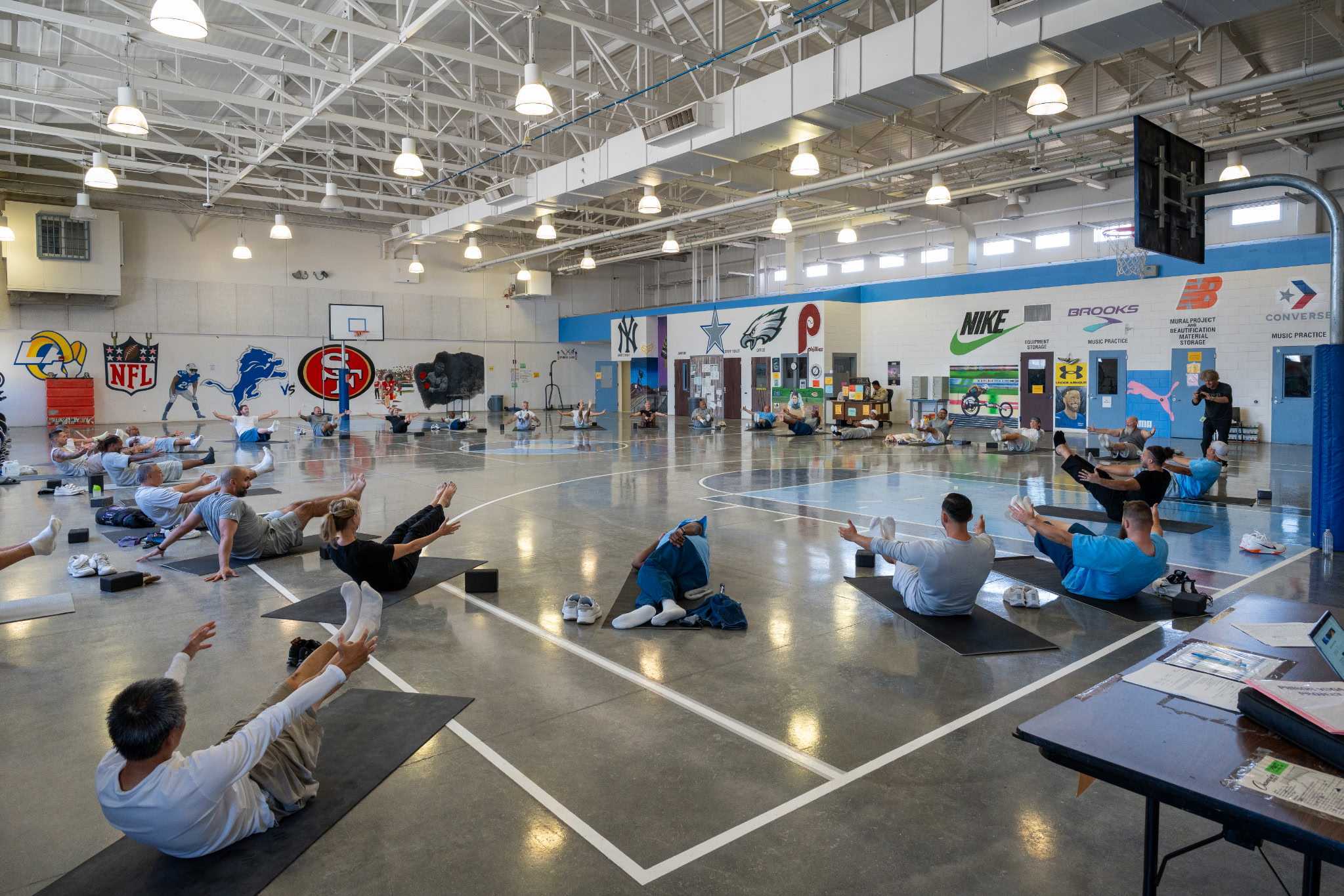 People exercising on mats in a large indoor sports facility with sports logos and equipment, bright lighting, and high ceilings.