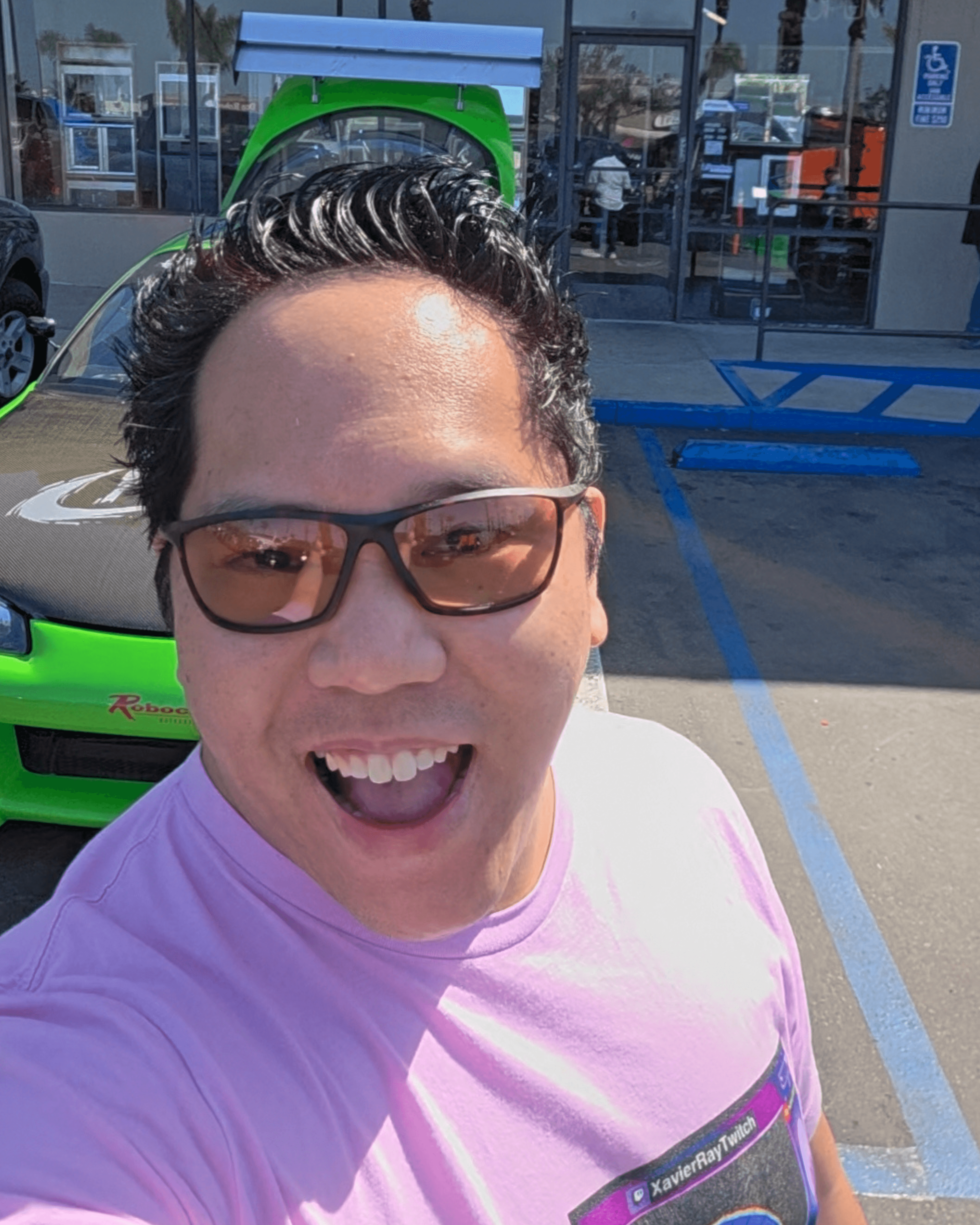 Person with glasses smiling outdoors in a parking lot, wearing a pink shirt, with a green vehicle behind them.