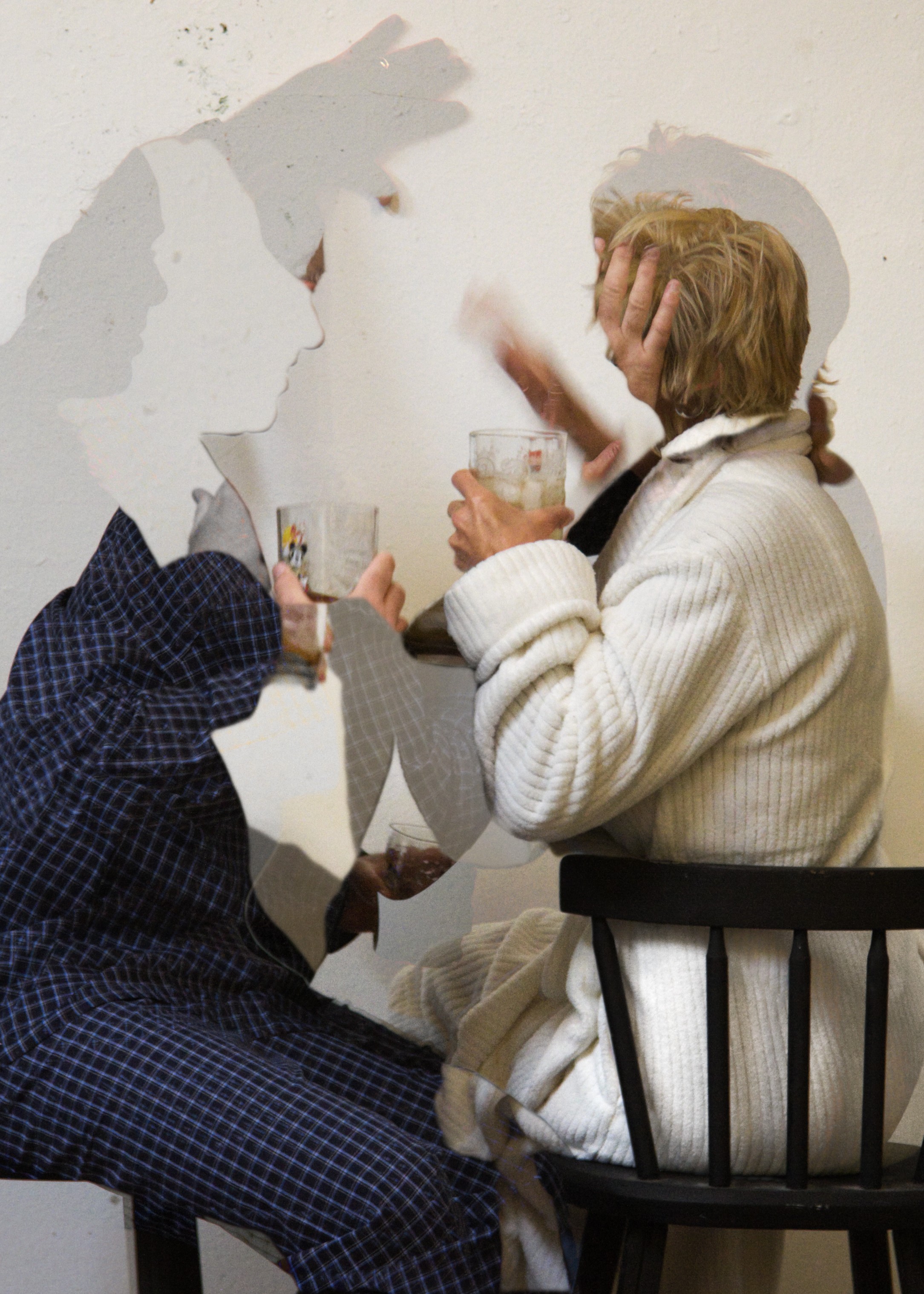 Two women with glasses of drink talking, shadows cast on a wall, one seated on a chair, the other standing.
