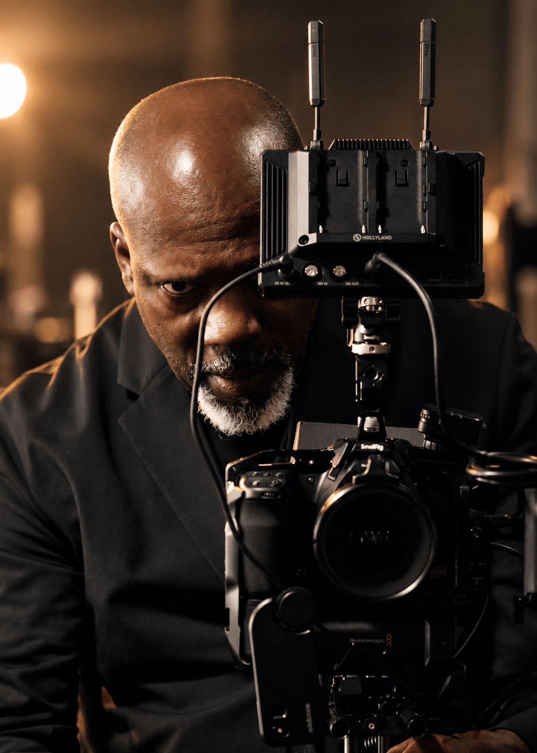 Man with a shaved head and beard operating a professional camera with wireless transmitters, in a dimly lit setting.