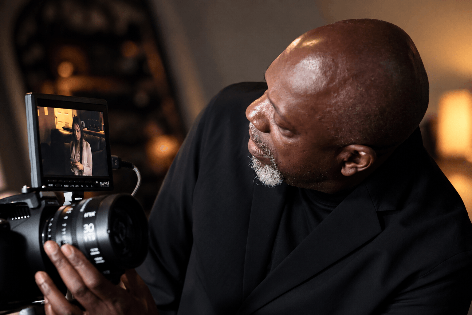Man with a shaved head and gray beard looks at a camera screen, indoors with warm lighting