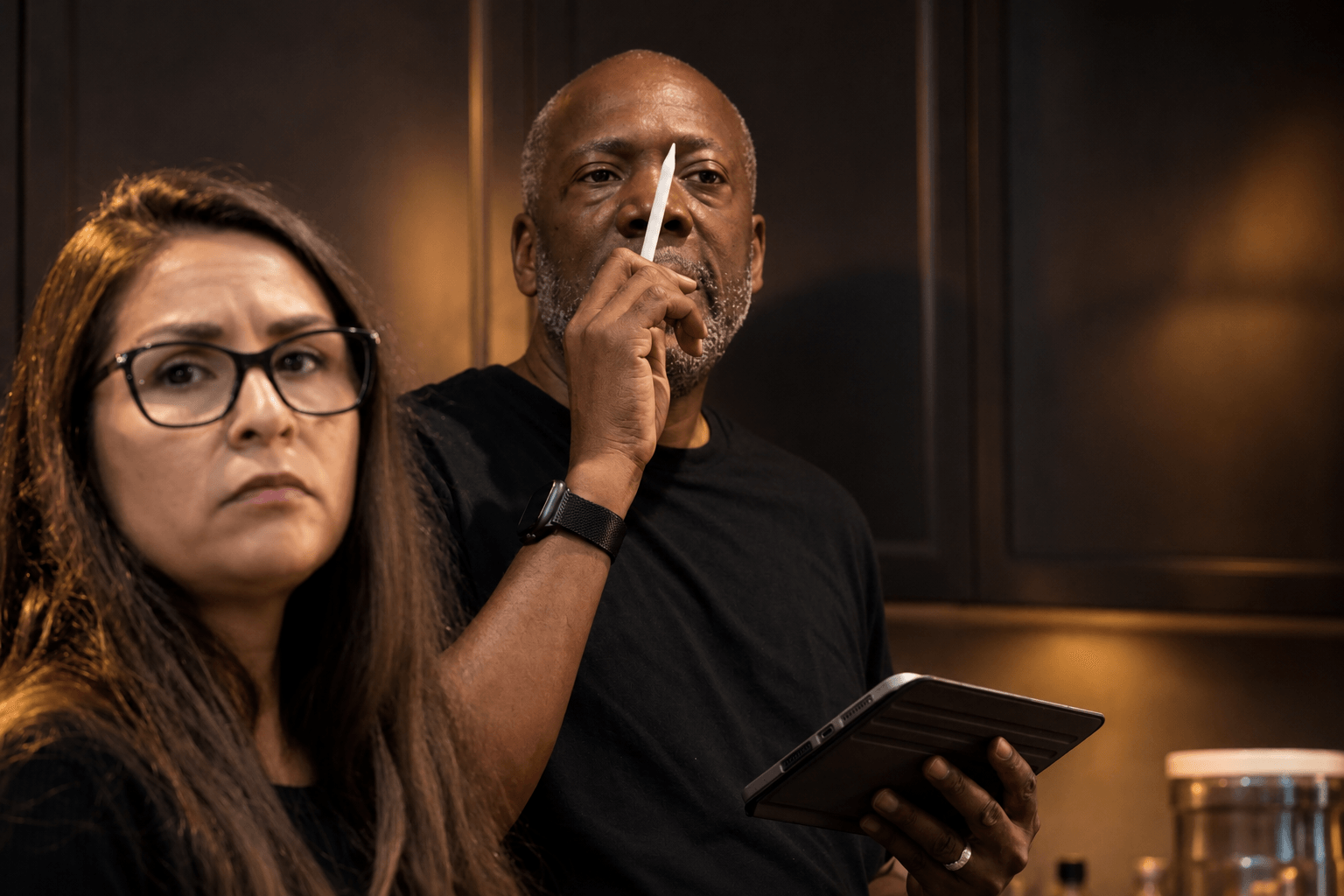 Two people, a woman with glasses and a man, in an indoor setting, with serious expressions, the man holding a tablet.