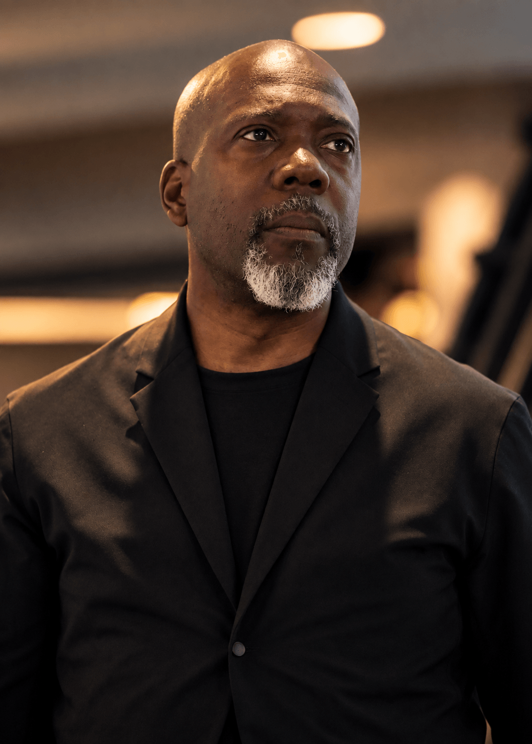 Man with a bald head and gray beard wearing a black blazer, looking to the side indoors.