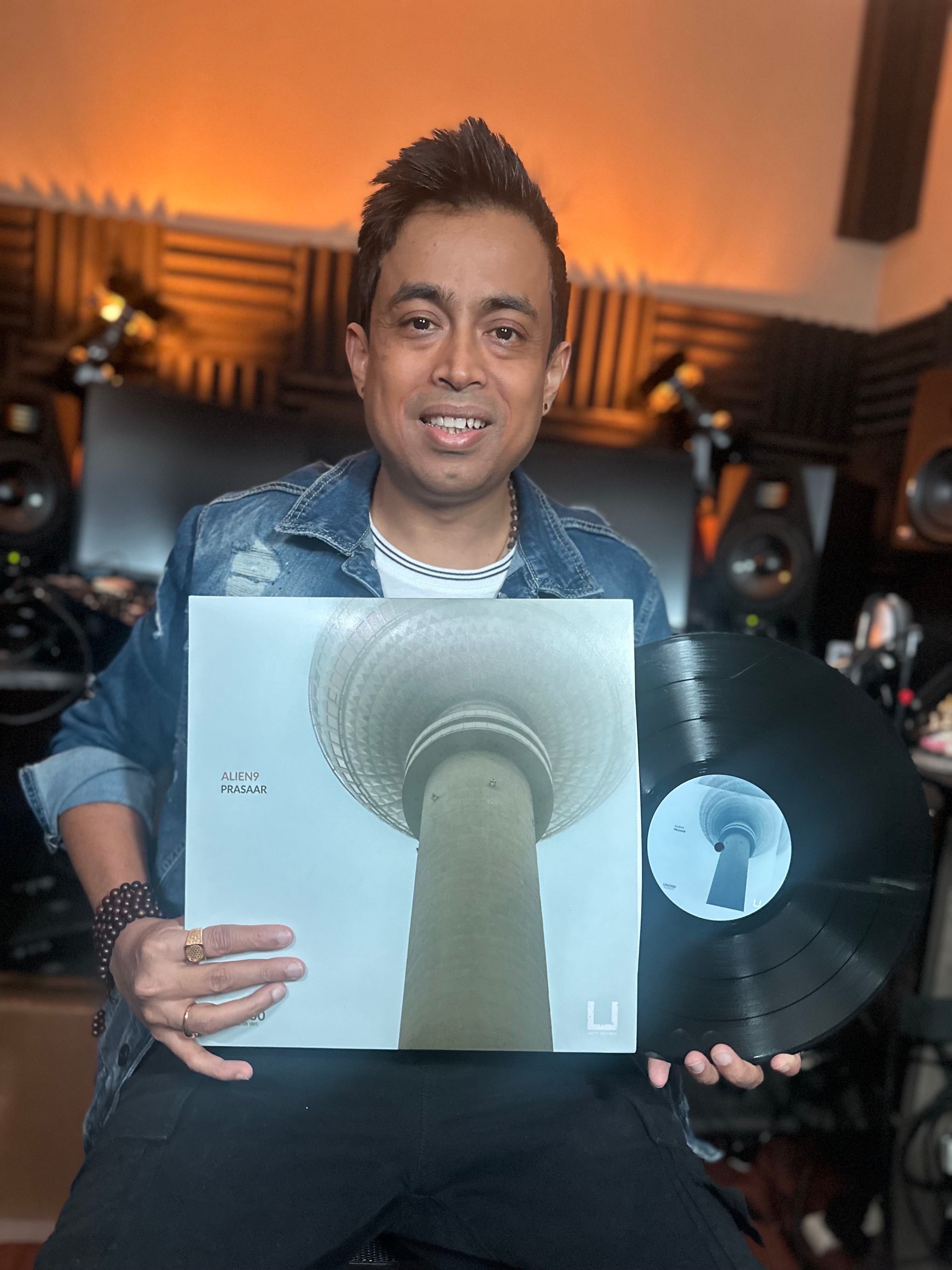 Person holding a vinyl record and album cover in a music studio with speakers and acoustic panels.