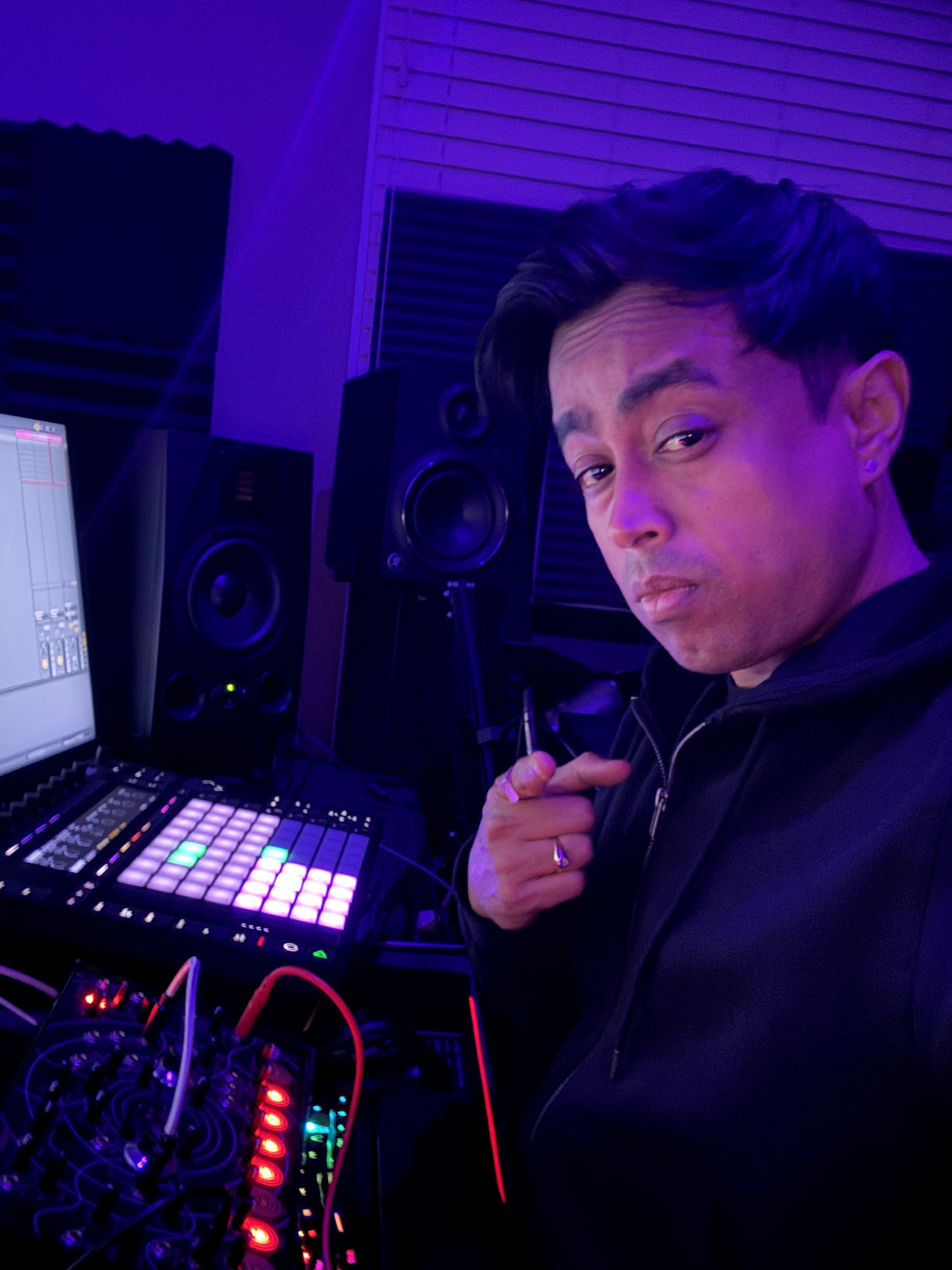 Young man in a music studio with audio equipment and speakers, holding a headphone cord, looking at the camera.