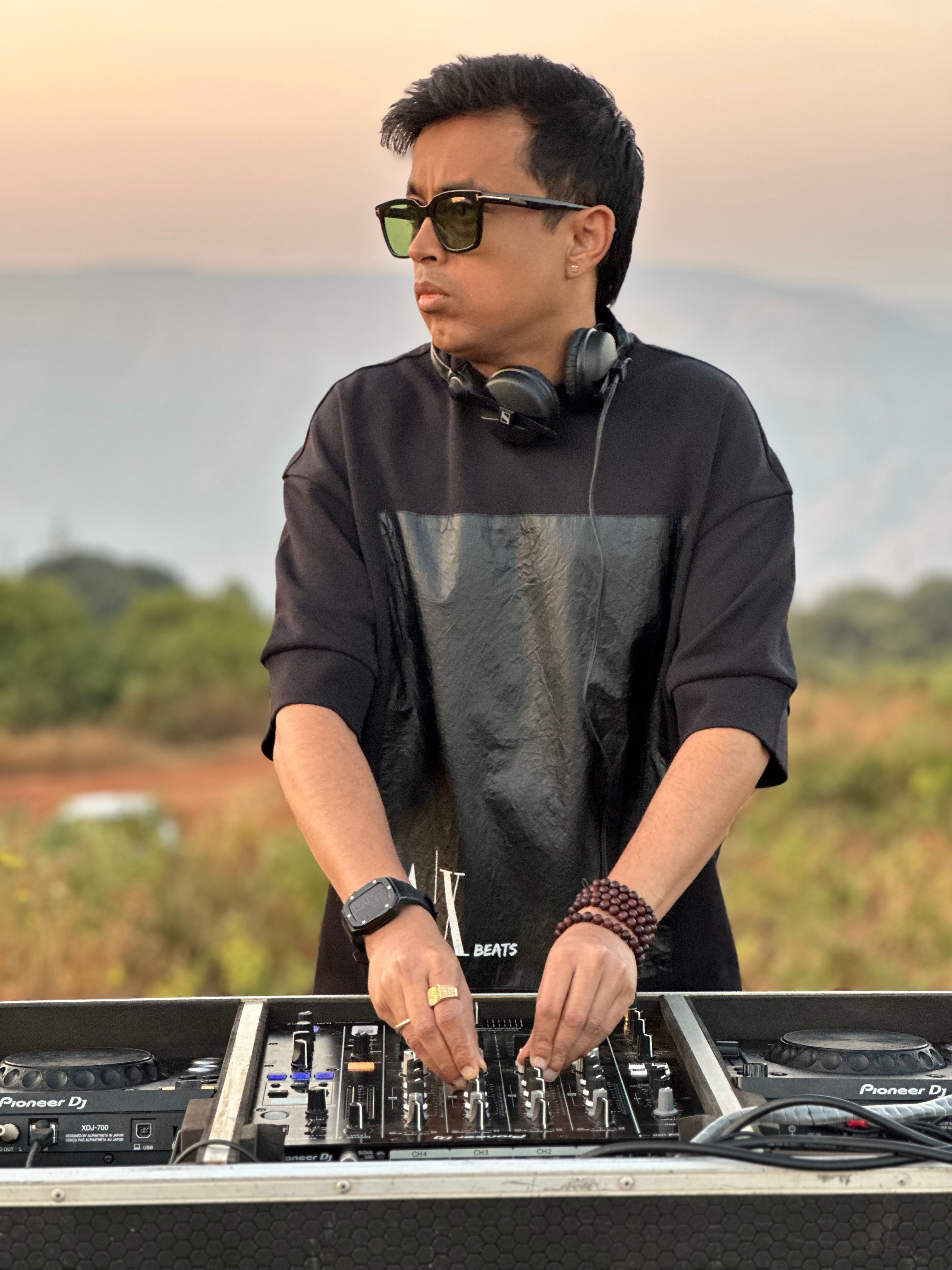 DJ wearing sunglasses and headphones, standing outdoors with a landscape background, operating a DJ mixer.