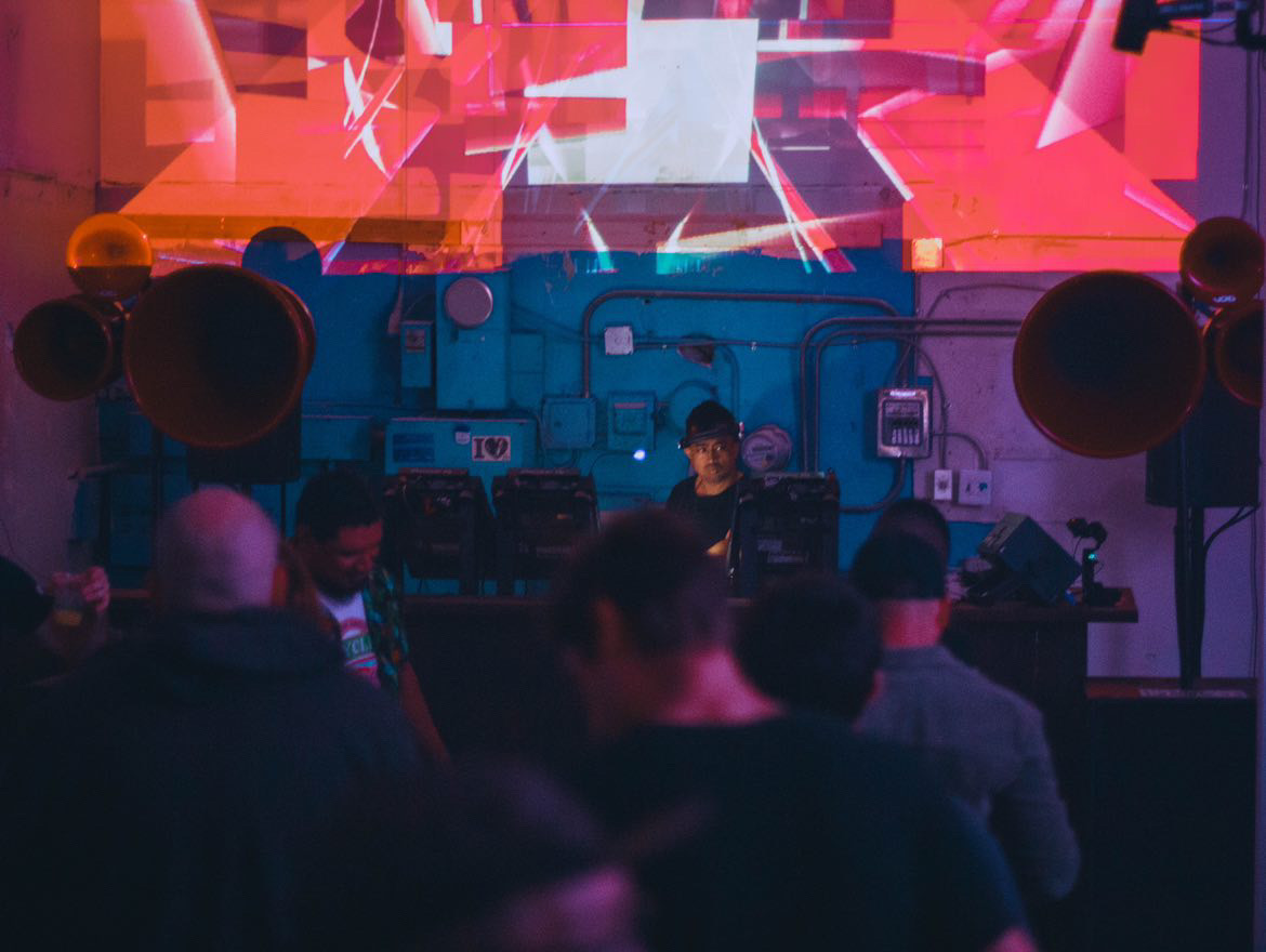 People watching a DJ perform on stage with colorful digital background, large speakers, and equipment, in a dark indoor setting.