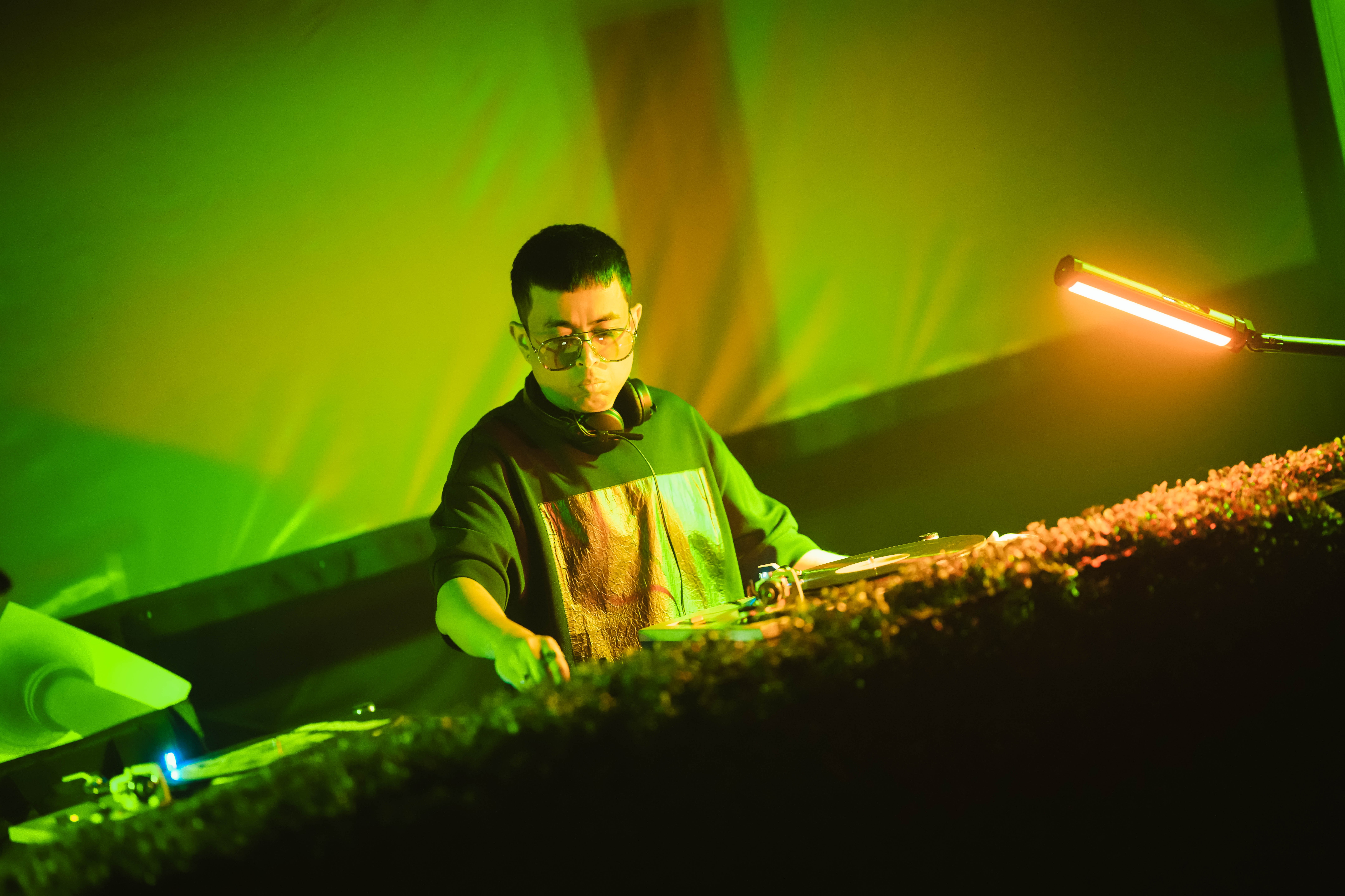 DJ wearing glasses and headphones, standing behind a mixing console with green and yellow lighting, focused on equipment.