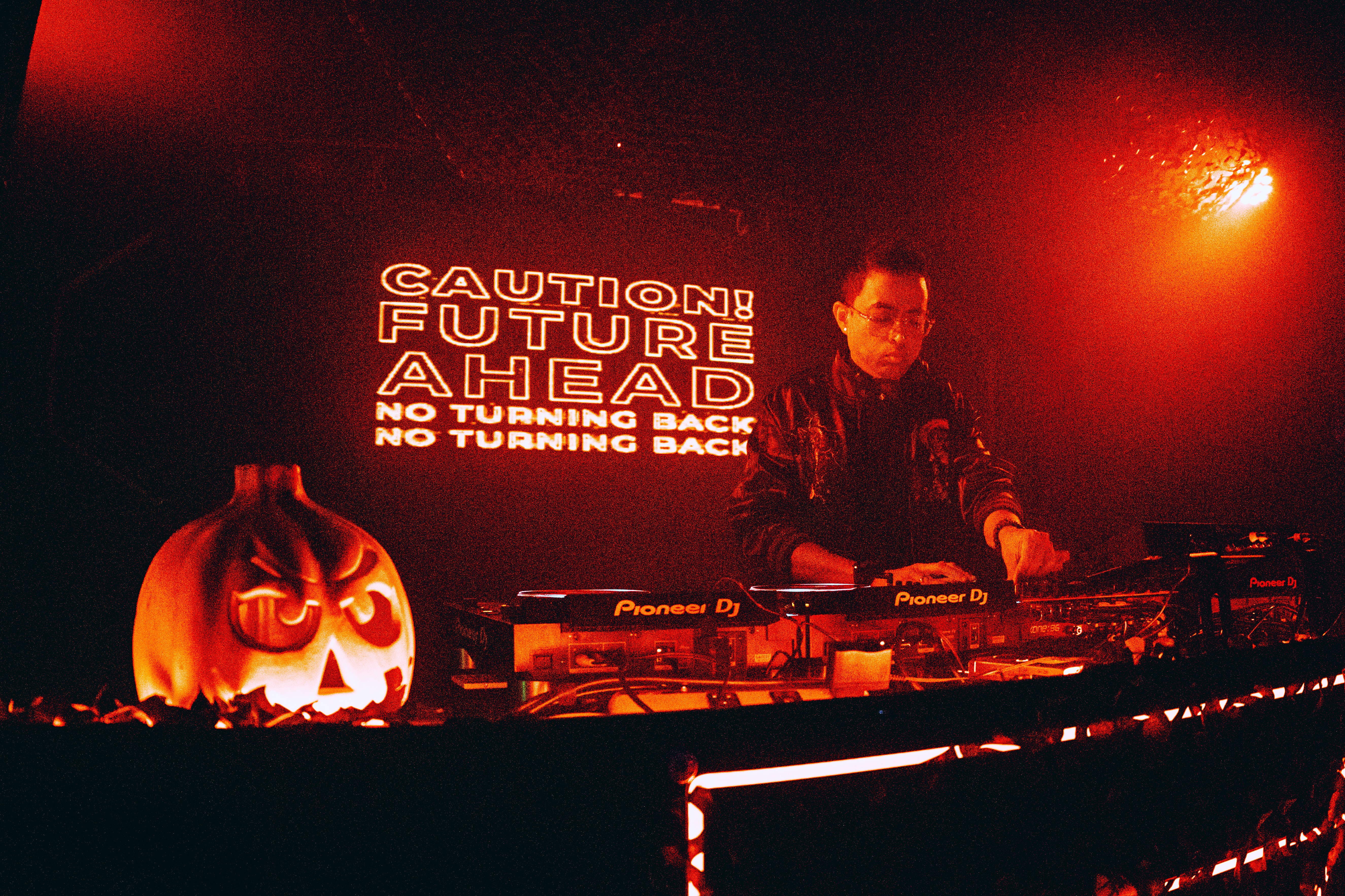 DJ performing with a carved pumpkin on the left, illuminated sign behind with caution message, dark background, red lighting.