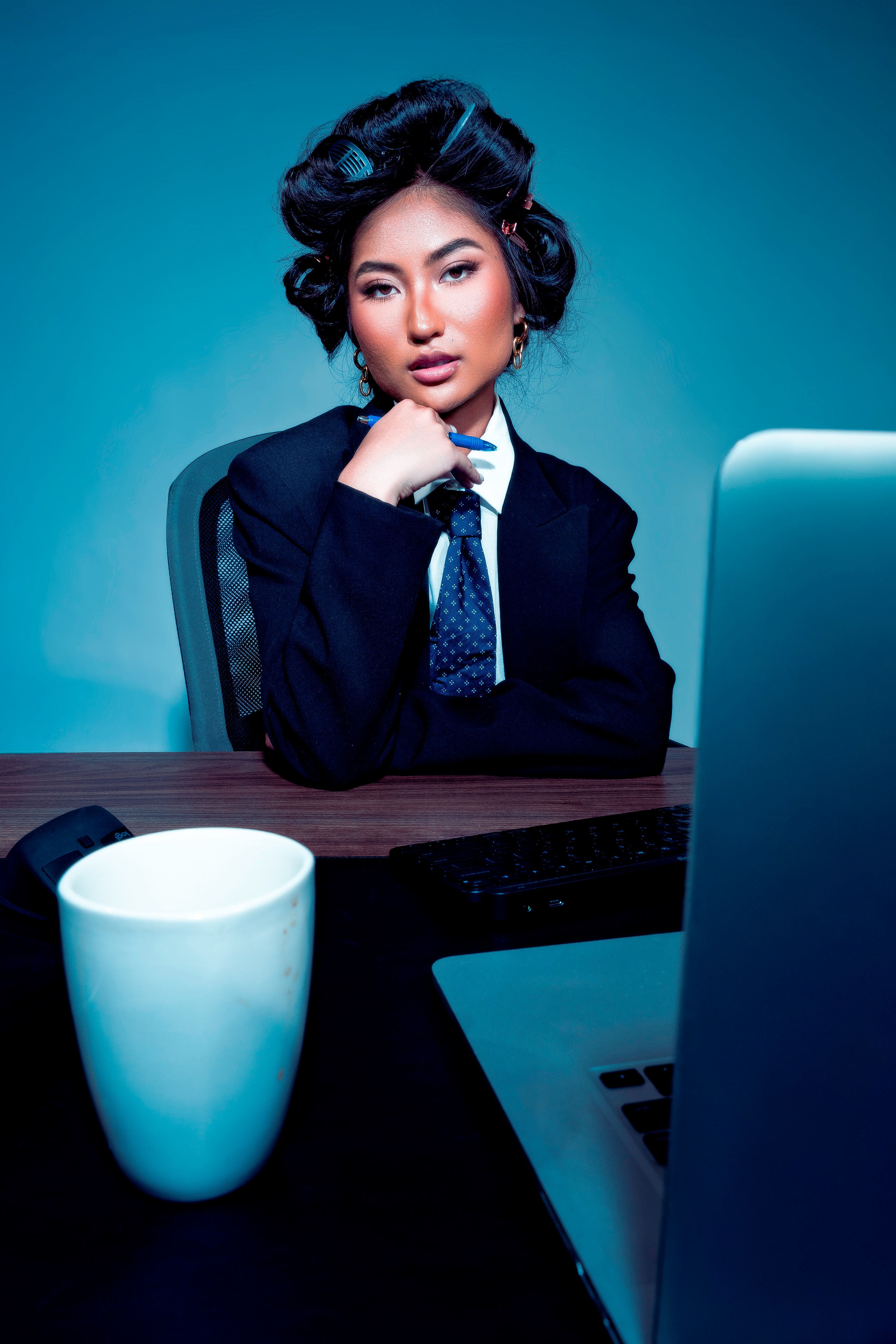 Woman in formal attire sitting at desk with laptop and coffee mug, looking at camera, blue background.