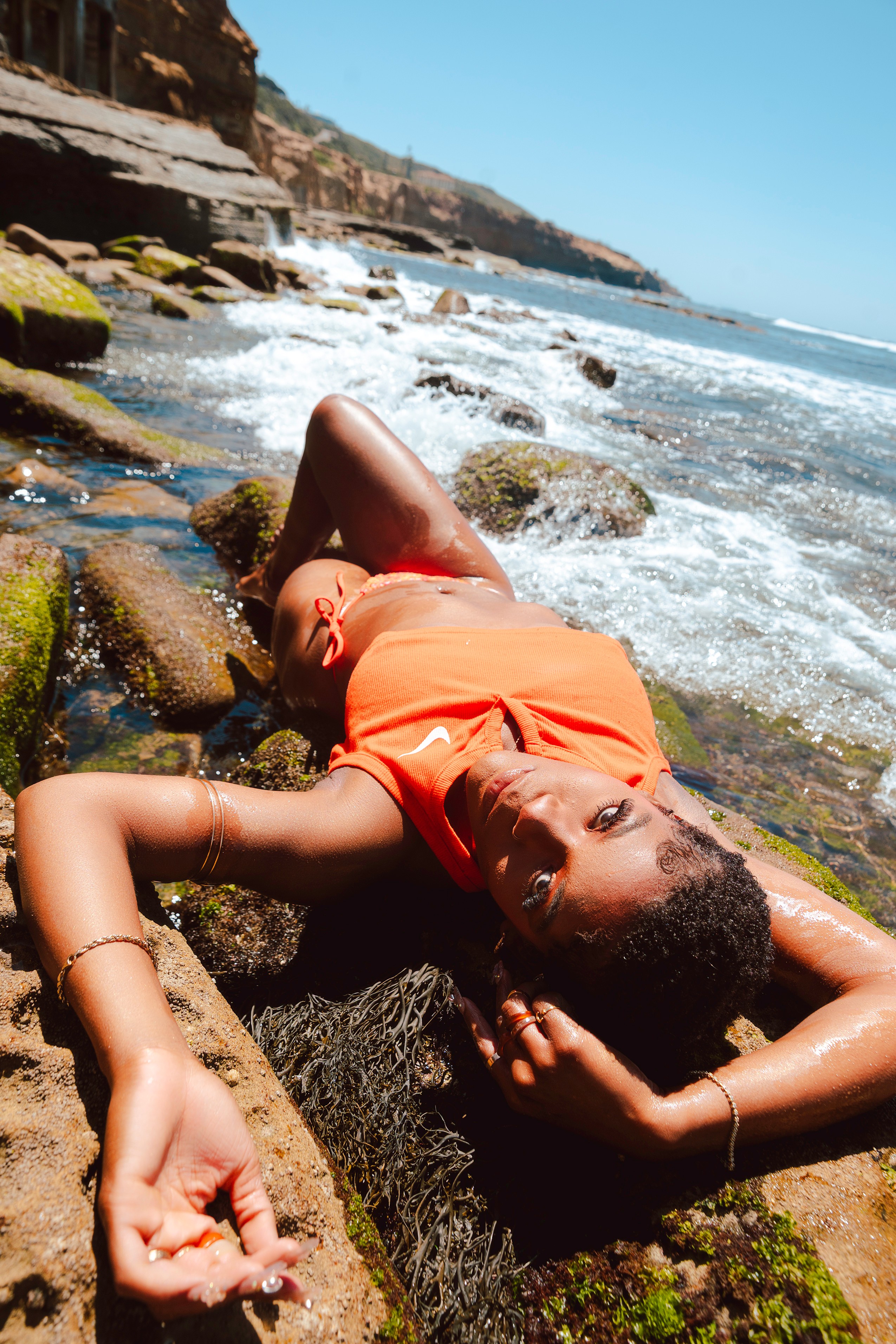 Woman lying on rocks by the ocean, wearing an orange top, with waves and cliffs in the background.