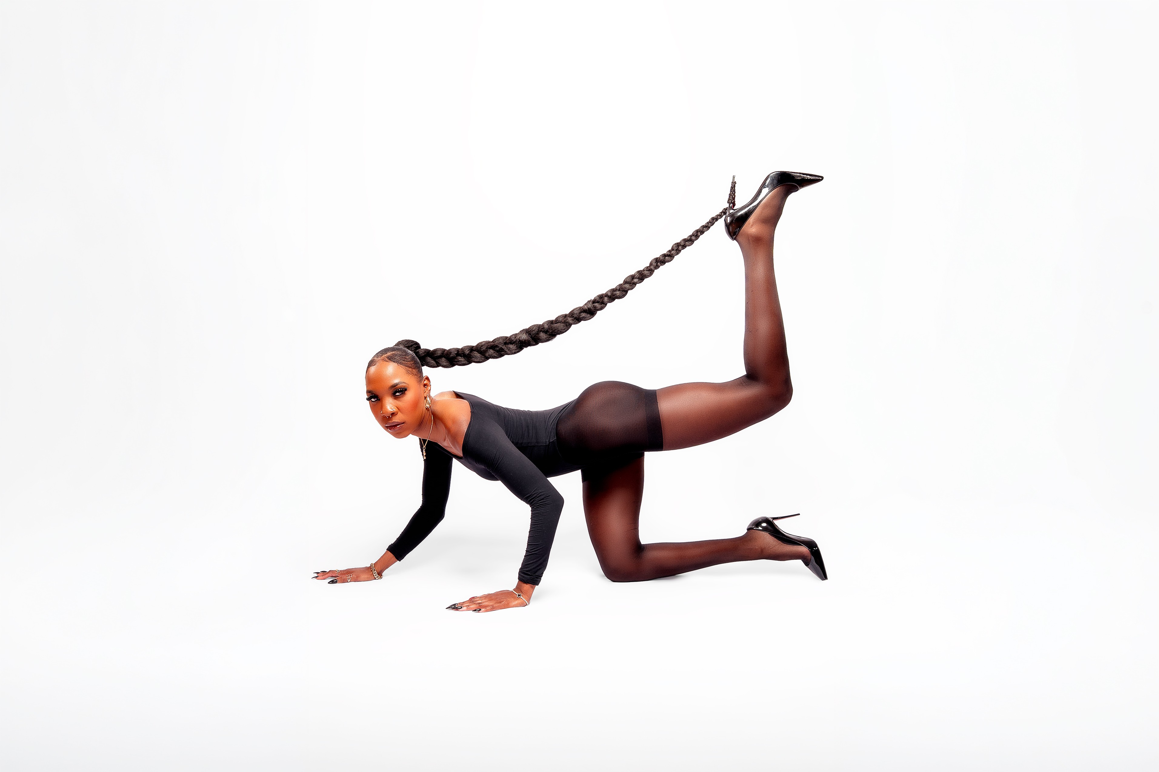 Person in black outfit with long braided hair, on hands and knees, against white background.