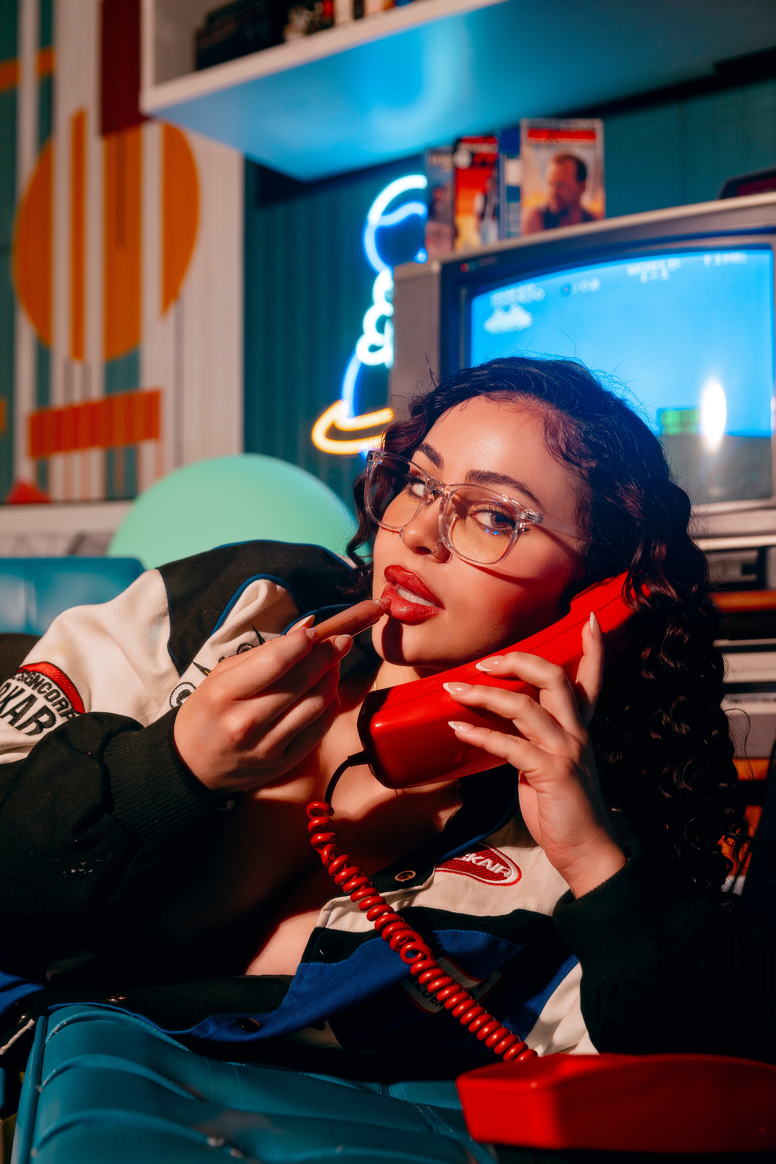 Woman with glasses talking on a red phone, holding lipstick, in a retro-themed room with neon lights and a TV.