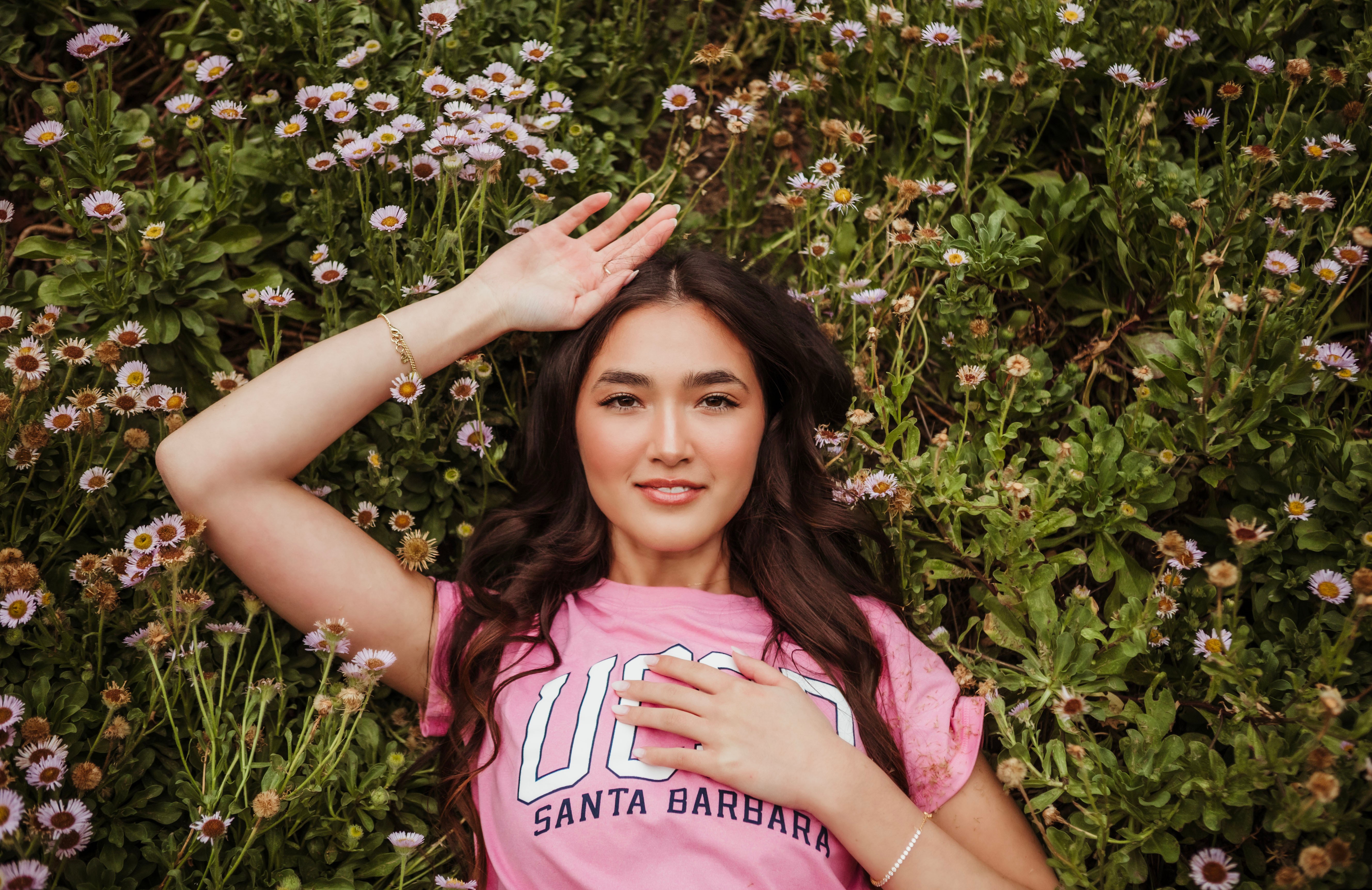 Young woman lying among flowers, smiling, with one hand on her forehead and the other on her chest, wearing a pink shirt.