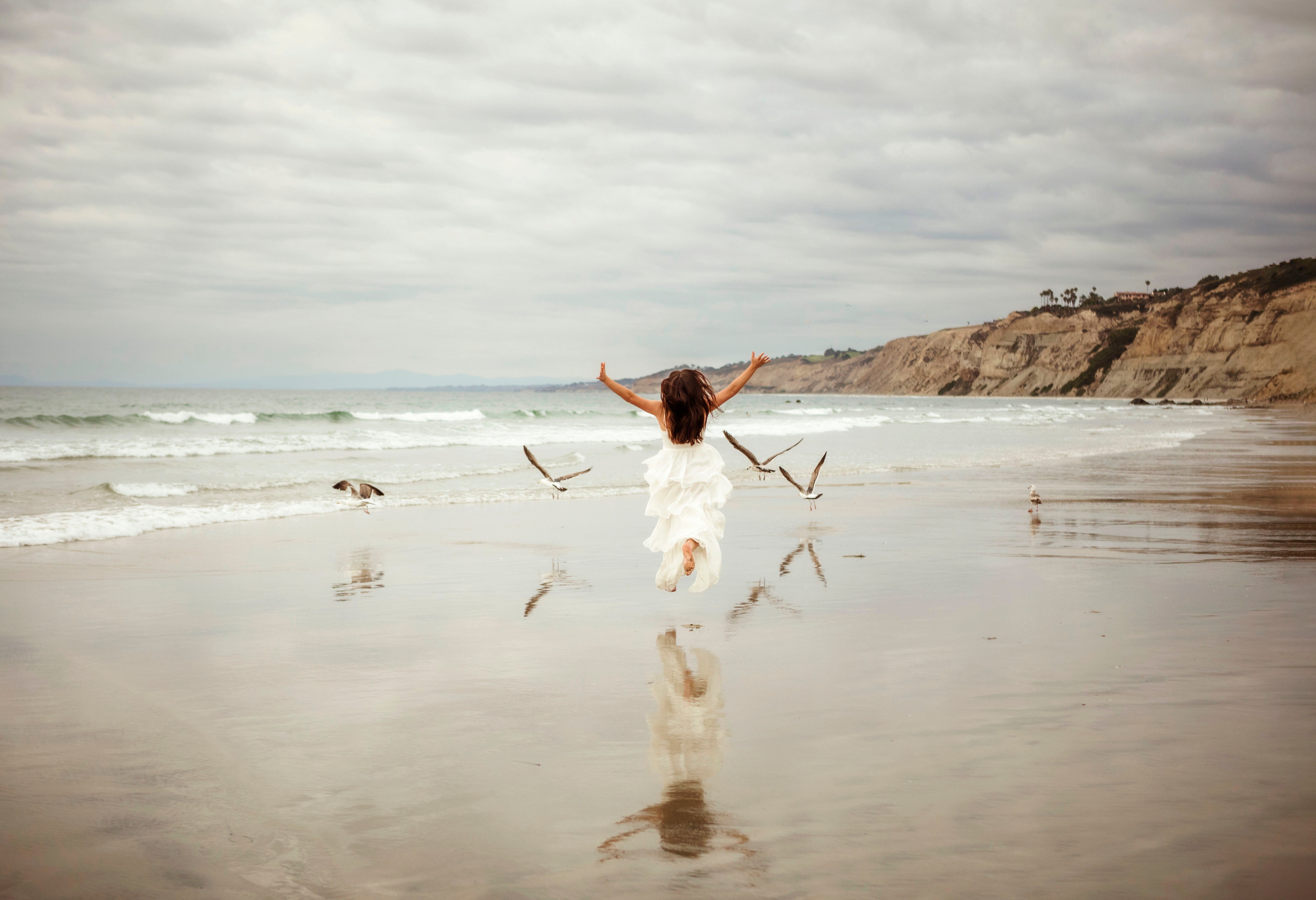 Person with arms raised on wet beach, ocean waves and cliffs in background, cloudy sky, reflection on wet sand.