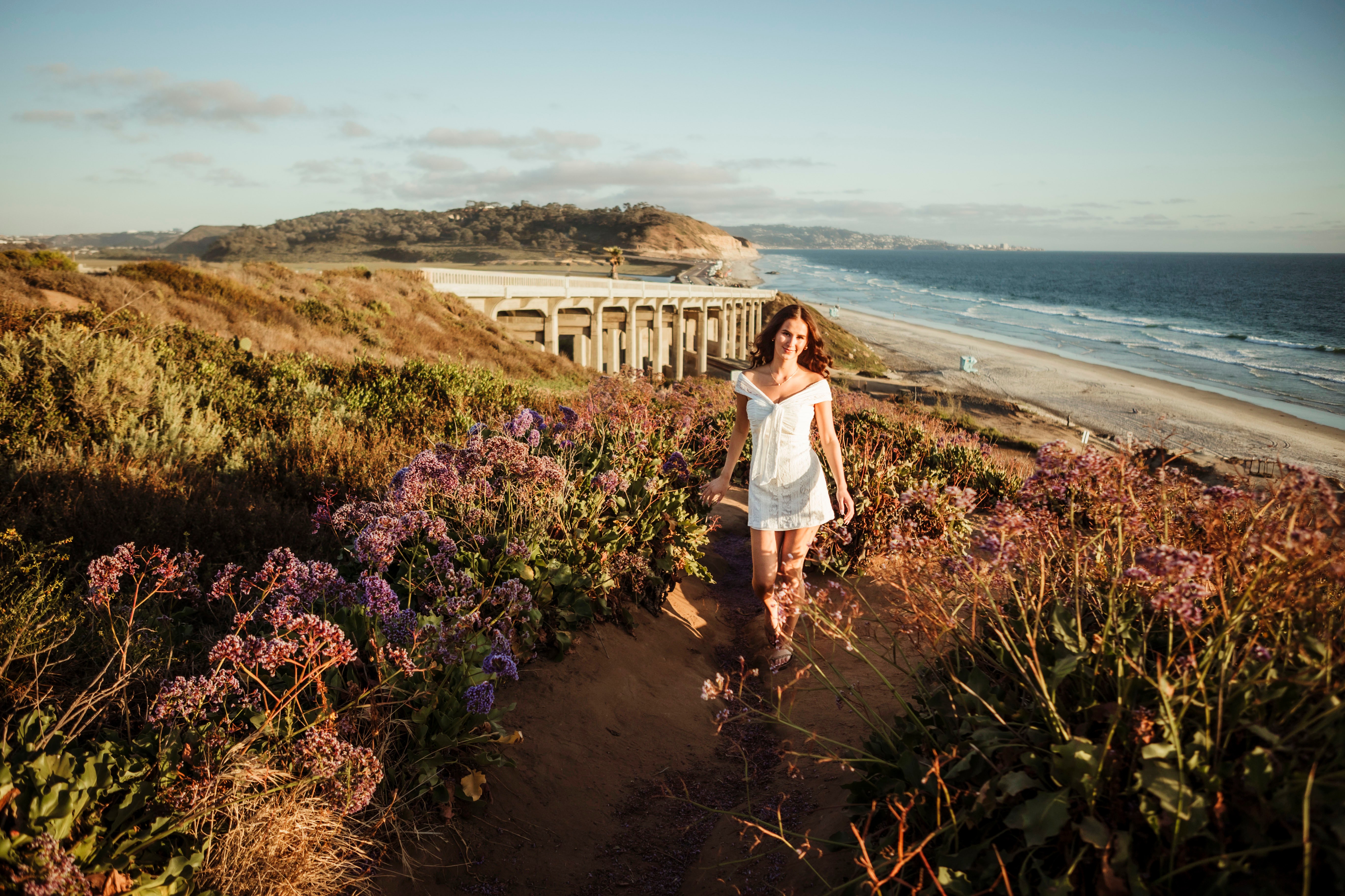 Woman walking along a coastal trail with purple and green plants, beach, and ocean in background, sunny sky.