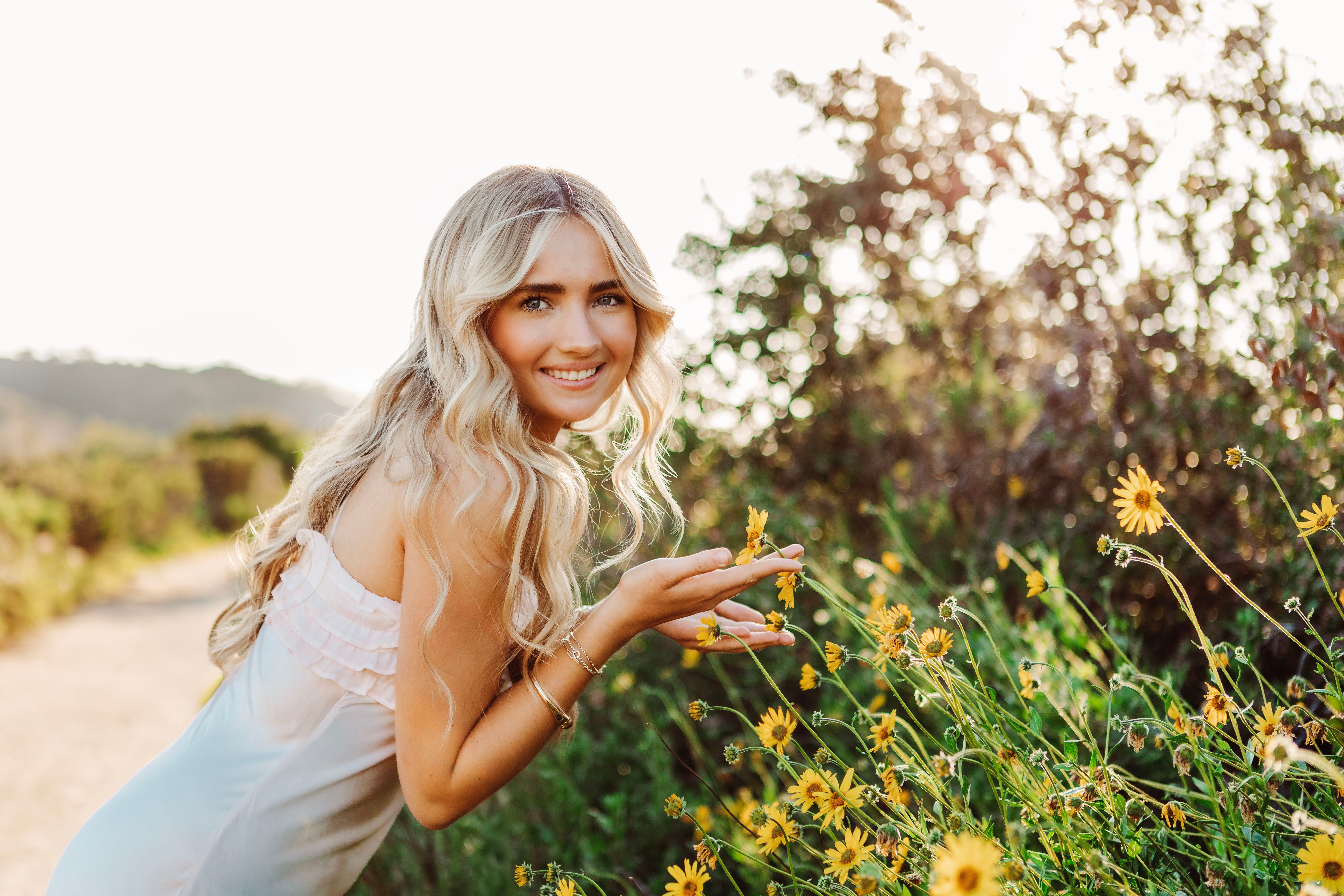 Smiling woman with long blonde hair in a white dress outdoors among yellow flowers, sunlight shining behind her.