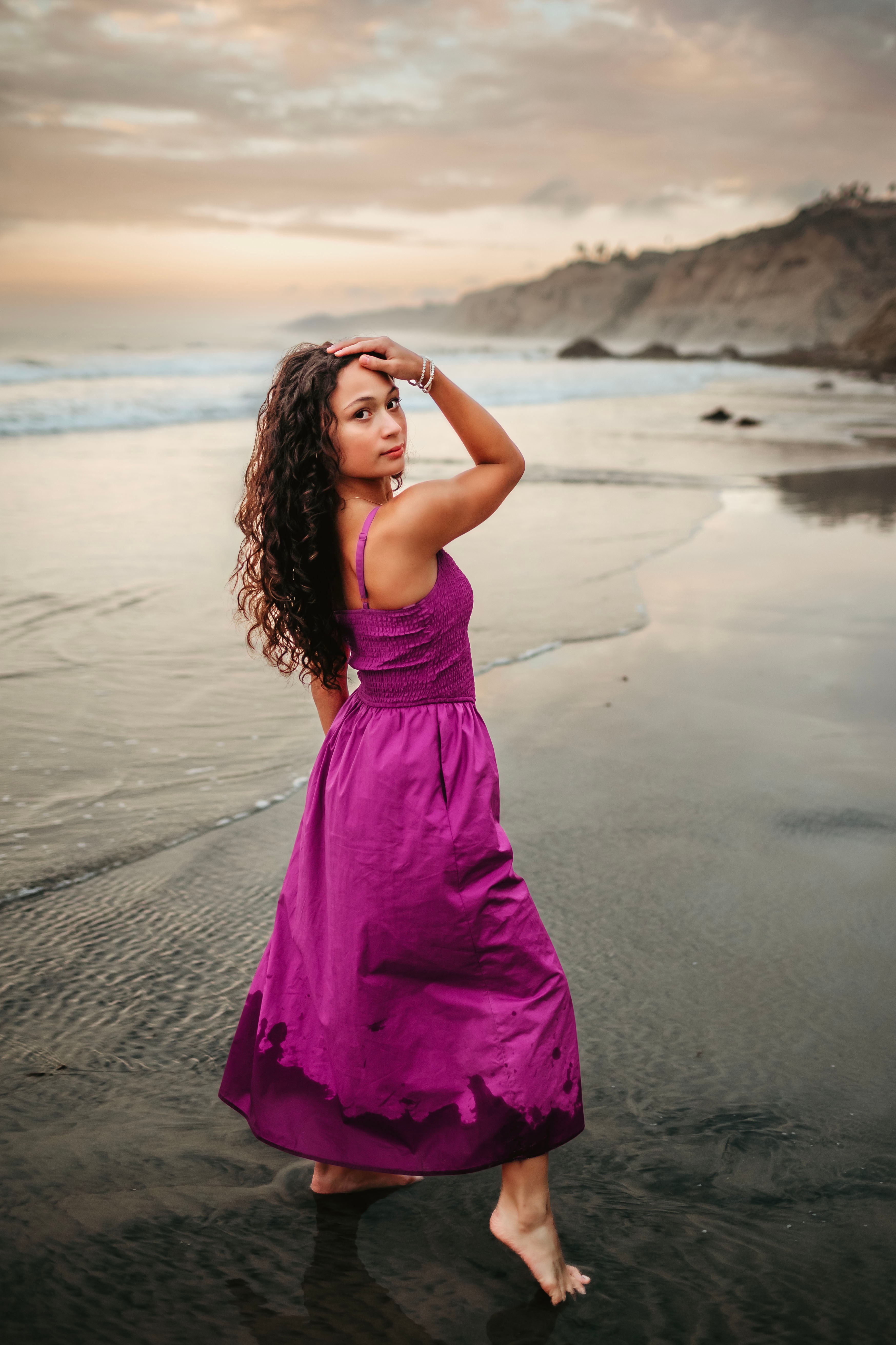Woman in a purple dress standing in shallow water on a beach with cliffs and cloudy sky in background.