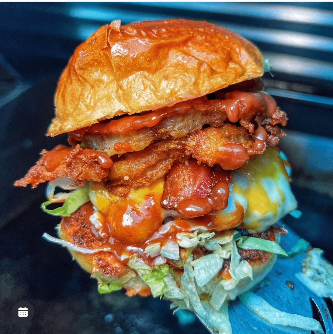 Close-up of a burger with bacon, lettuce, tomato, and sauce in a bun, on a dark background.