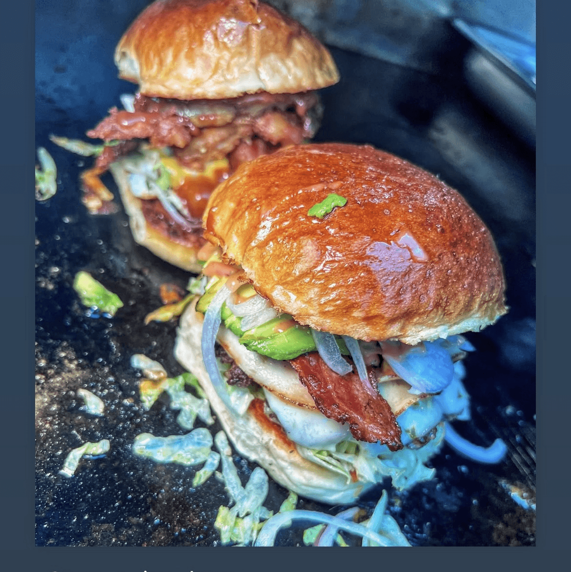 Two burgers with buns, lettuce, tomato, and meat, on a black surface with crumbs and shredded lettuce.
