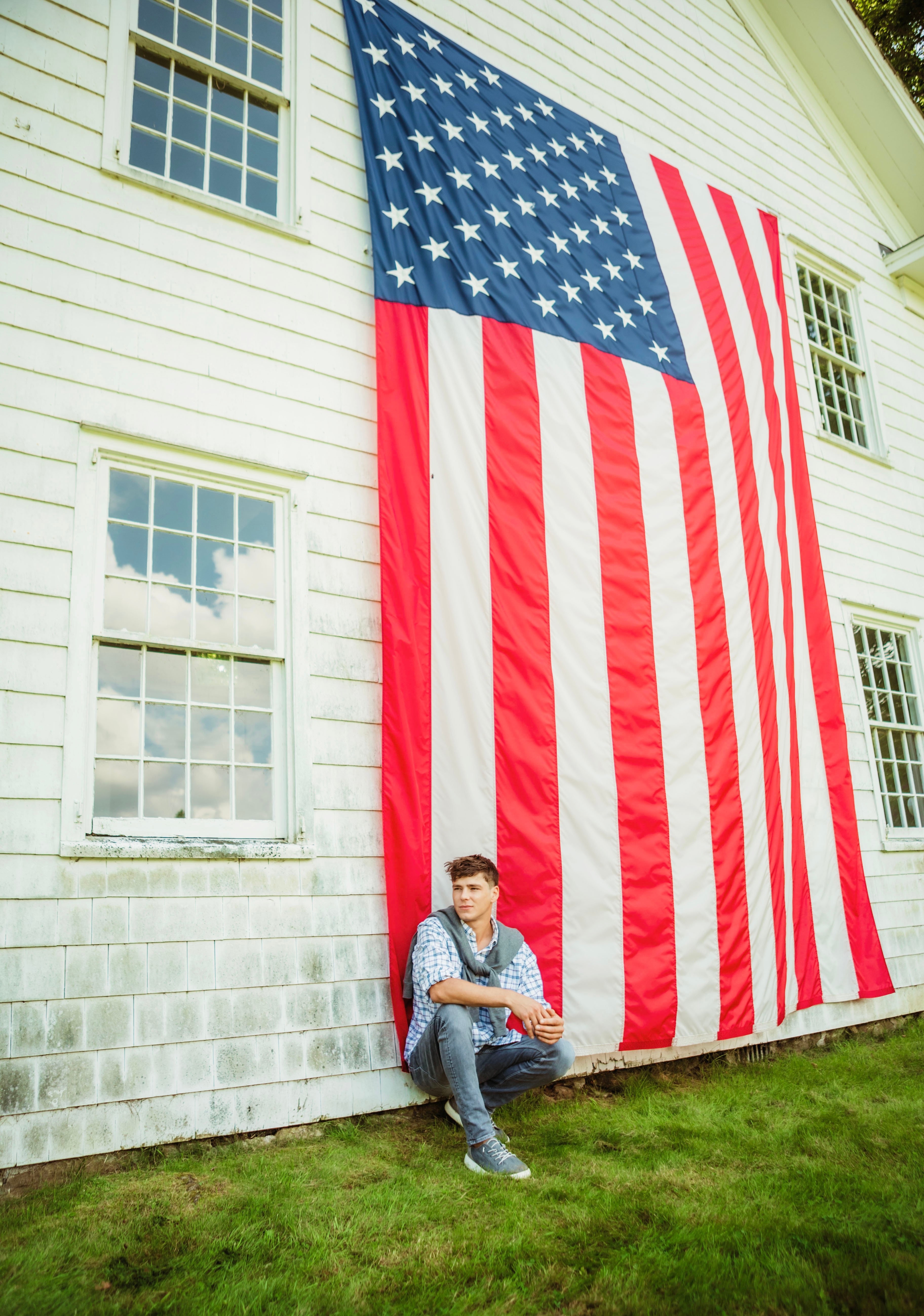 Boy leaning against a white house with large American flag hanging on the wall, grass in foreground.